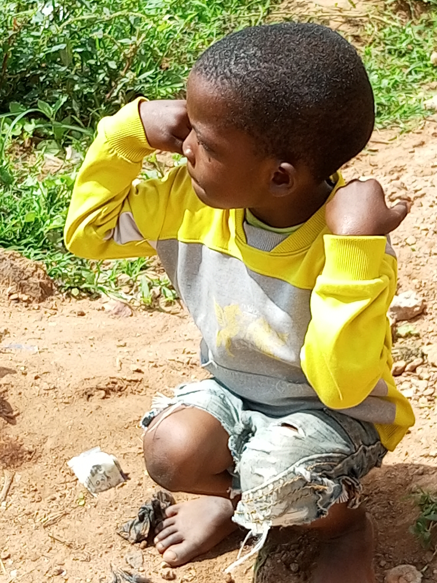 A young child with dark skin and short, curly black hair is kneeling on sandy ground. The child is wearing a yellow long-sleeved shirt with a grey panel across the chest, and ripped denim shorts. The child's right arm is raised, and their hand is near their forehead, as if shielding their eyes or in thought. The background consists of green foliage and dry, brown earth. The scene appears to be outdoors in a rural or semi-rural setting. The child is the sole subject of the image.