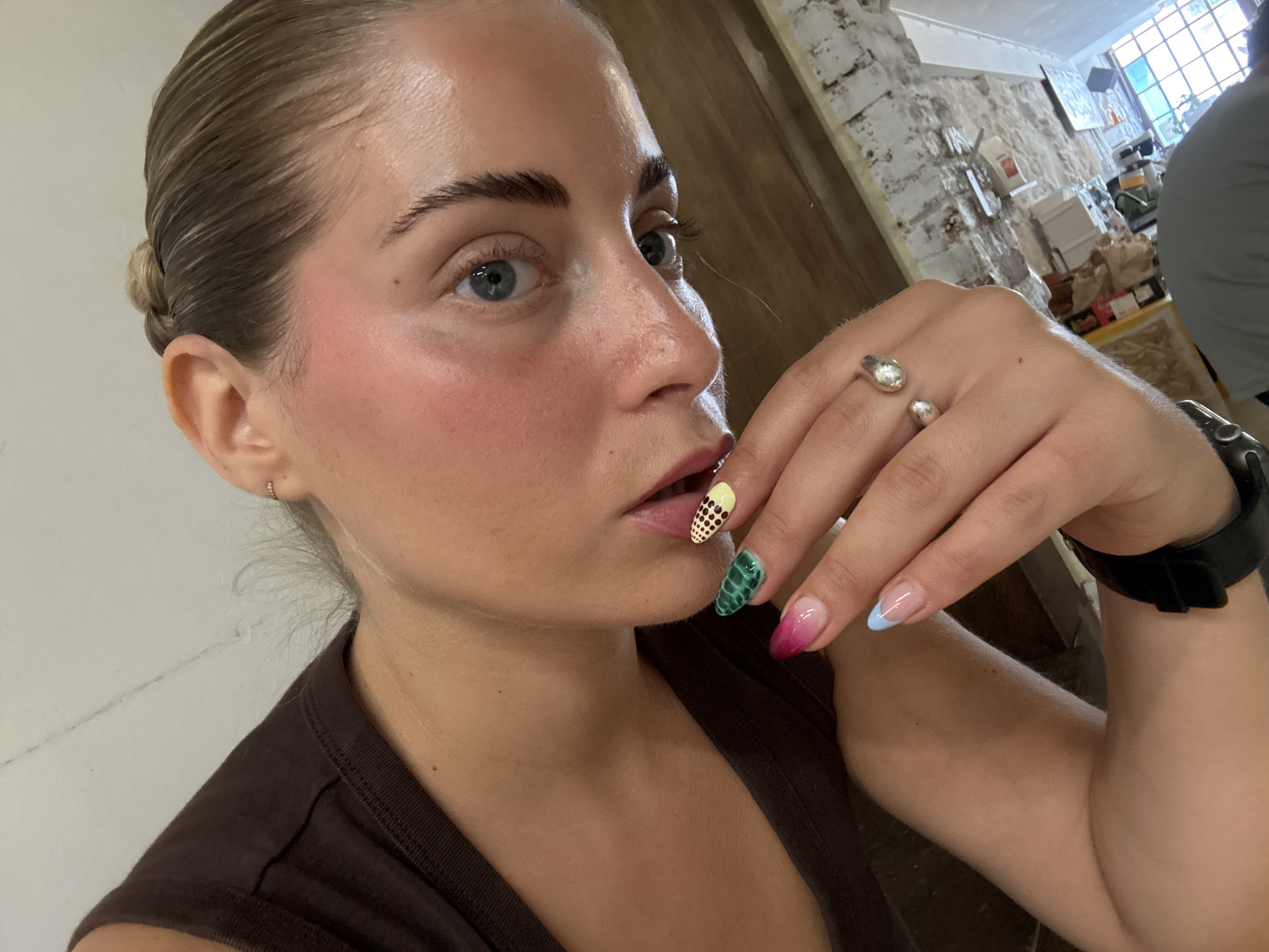 The image shows a close-up, overhead shot of a young woman's face and upper body. She has blonde hair pulled back into a bun and blue eyes. She is wearing makeup, including blush, and has on a brown top. Her left hand is near her face, showing off her brightly painted fingernails, which feature various designs including dots, green scales, and pink and blue French tips. She is also wearing a silver ring and a black watch on her left wrist. The background appears to be an indoor setting, possibly a cafe or restaurant, with a brick wall and some shelves with items on them visible. The lighting suggests daytime. There is no indication of the location being Paris, France.
