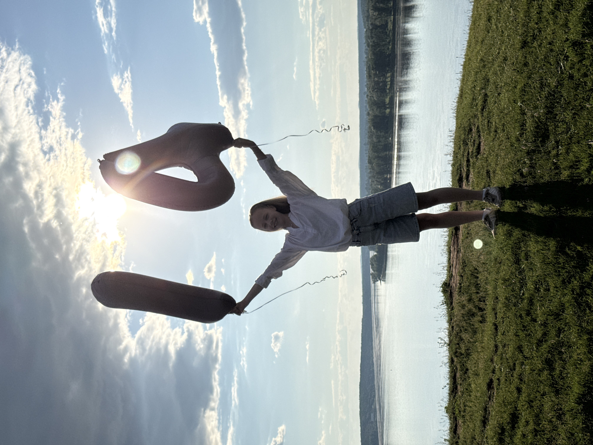 A full-body, outdoor photograph depicts a female subject, estimated to be a teenager, standing centrally on a grassy knoll overlooking a river in Ольховка, Russia. The subject, with light-colored hair, is observed smiling towards the camera. She wears a white long-sleeved collared shirt, light-wash denim shorts, and light-colored athletic shoes. She holds two large, gray, deflated or partially deflated foil balloons above her head: one shaped like the numeral '1' is held in her left hand, and one shaped like the numeral '4' is held in her right hand. Both balloons have attached strings. The sun is positioned directly behind the subject and the '4' balloon, generating significant backlighting and lens flare in the upper right quadrant. The sky above features a bright sun partially obscured by white and gray clouds against a blue backdrop. Below the horizon, a wide, calm river extends, bordered by a dense line of green trees on the distant bank. The immediate foreground is a green, textured grassy slope with visible soil patches. The subject's shadow is cast forward onto the grass.
