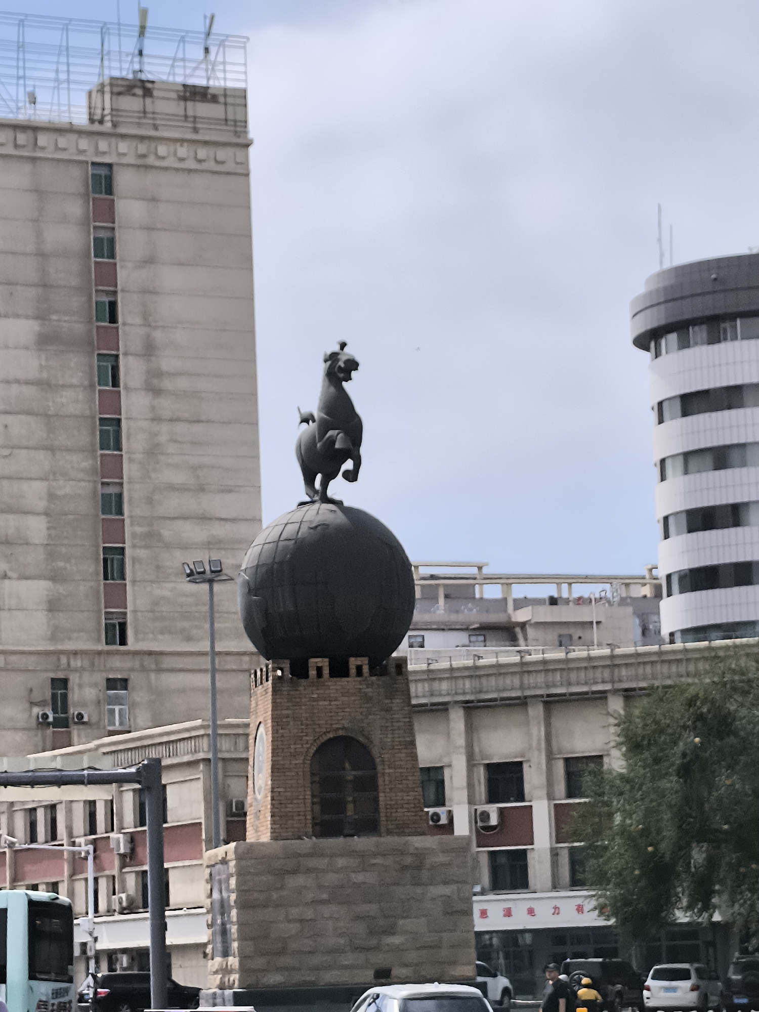 The image shows a statue of a horse standing on a globe, which is mounted on a brick pedestal. The statue is located in a city, with tall buildings visible in the background. To the left of the statue is a tall, light-colored building with many windows. To the right of the statue is a rounded, modern building with white horizontal bands. The sky is overcast. In the foreground, there is a street with a bus, cars, and some pedestrians. There is also a tree with green foliage. The statue is likely located in Ürümqi, China, as suggested by the context.