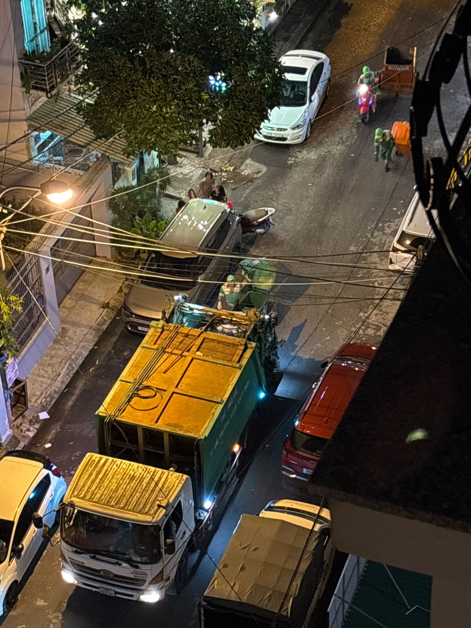 The image captures a nighttime scene on a street in Nha Trang, Vietnam. A garbage truck with a yellow and green body is parked on the right side of the street, illuminated by its headlights and a blue light on its side. In front of the garbage truck is a white truck, its headlights also on. To the left of the white truck is a white car. Further down the street, a red SUV is parked, and beyond that, a white sedan is visible. Several people are present in the scene, including some on the sidewalk and others near a motorbike. Two individuals in green uniforms are operating a motorbike and pushing a garbage bin, suggesting a waste collection operation. The street is lined with buildings and trees, and power lines crisscross overhead.