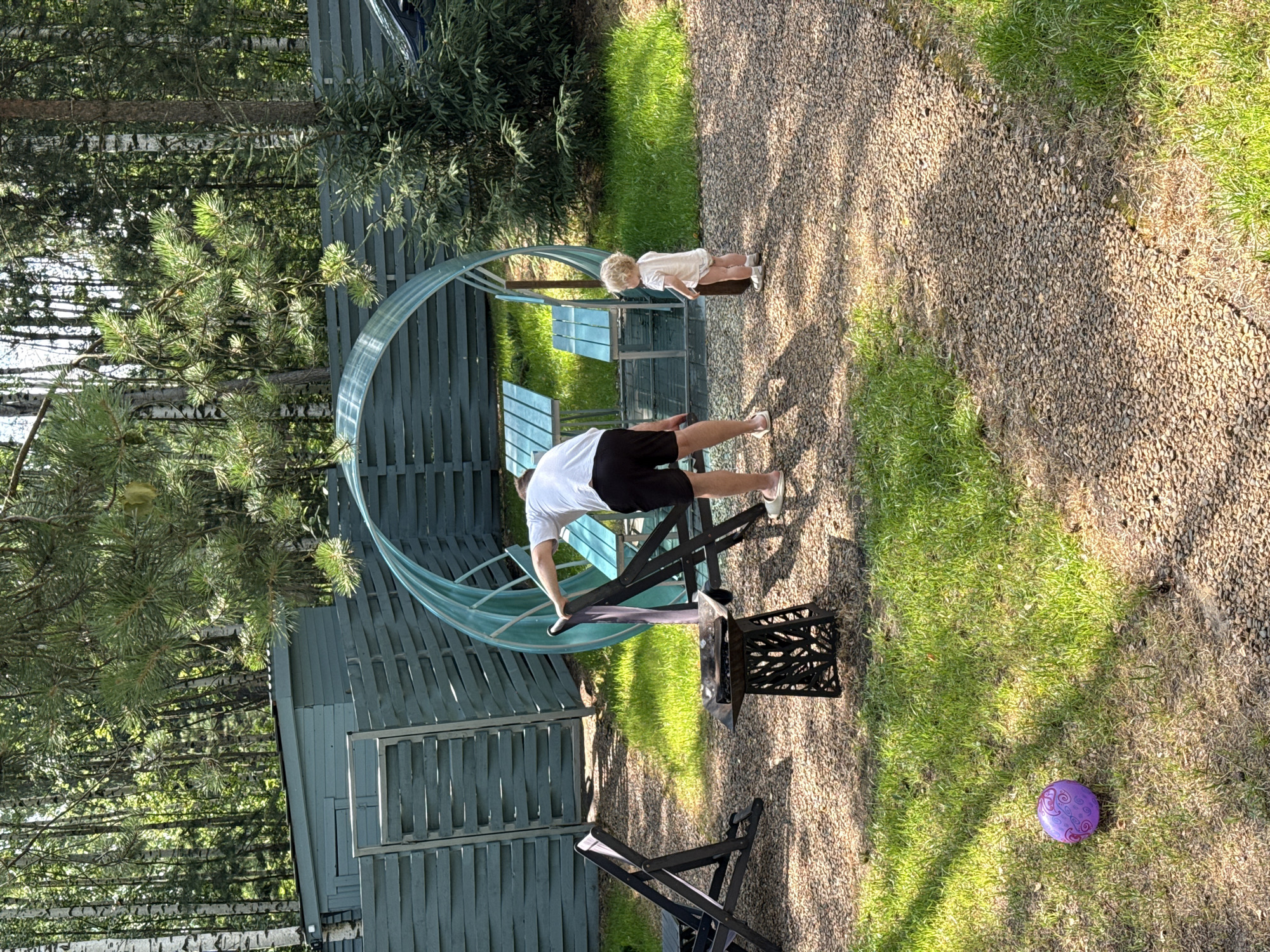 In Prokudino, Russia, a man in a white t-shirt and black shorts stands near a circular, translucent greenhouse structure with blue benches inside, adjusting a black folding chair. A toddler, wearing a light-colored onesie and diaper, stands to the right of the greenhouse, facing away from the camera. In the foreground, a purple ball rests on a patch of grass, with gravel paths and more grass surrounding the area. A dark metal brazier with a decorative base sits between two black folding chairs. The scene is set in a yard with a wooden fence and trees in the background.