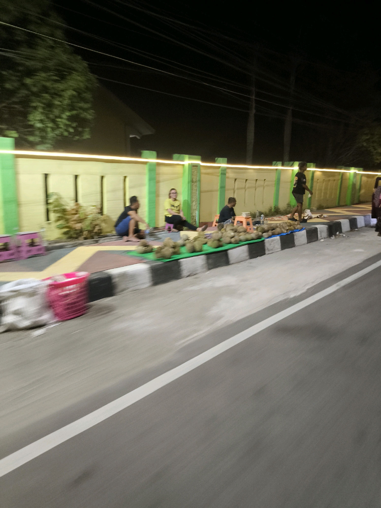 In Kwandang, Indonesia, at night, several people are visible near a roadside stall. One individual, wearing a black t-shirt and dark shorts, is walking away from the camera. Another person, seated on the ground, appears to be interacting with a bottle of water. A third person, wearing a yellow t-shirt and dark pants, is seated on a stool. A large pile of durians is displayed on a green mat in front of the seated individuals. The scene is illuminated by overhead lighting, casting a yellow glow along a decorative wall and green pillars. In the foreground, a pink basket and some white bags are visible on the ground next to a road with a white line marking. The overall impression is that of a nighttime marketplace or roadside vendor setting.