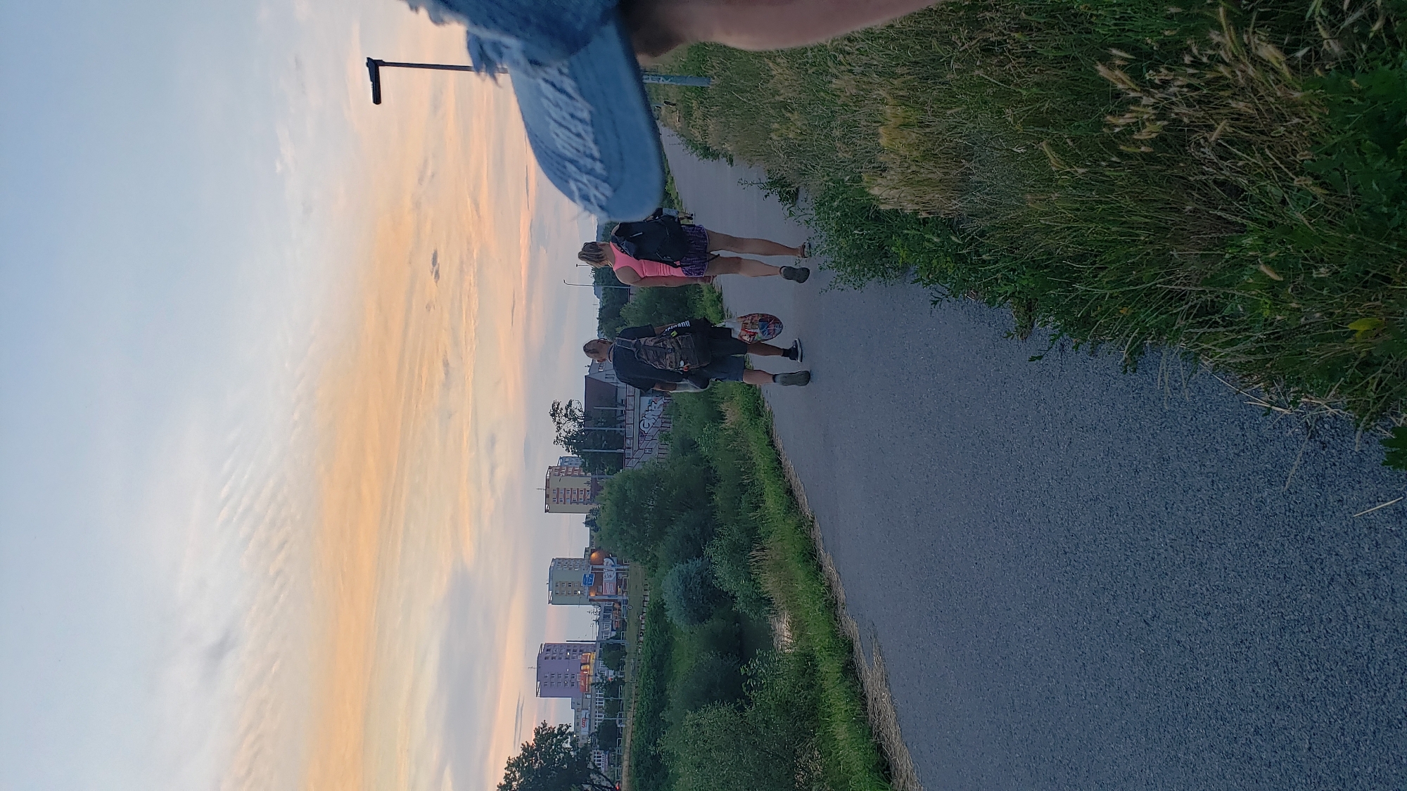 The image shows a paved pathway bordering a grassy area with a river on the left. Two individuals are walking away from the camera on the pathway, each carrying a backpack. The male in front is wearing a black t-shirt and shorts, and the female behind him is wearing a pink tank top and patterned shorts. In the background, several tall, multi-colored buildings are visible, characteristic of the Sekčov housing estate in Prešov, Slovakia. The sky is a soft gradient of light blue and orange, suggesting it is either dawn or dusk. The foreground shows the edge of a blue cap, suggesting the photo was taken by someone wearing it.