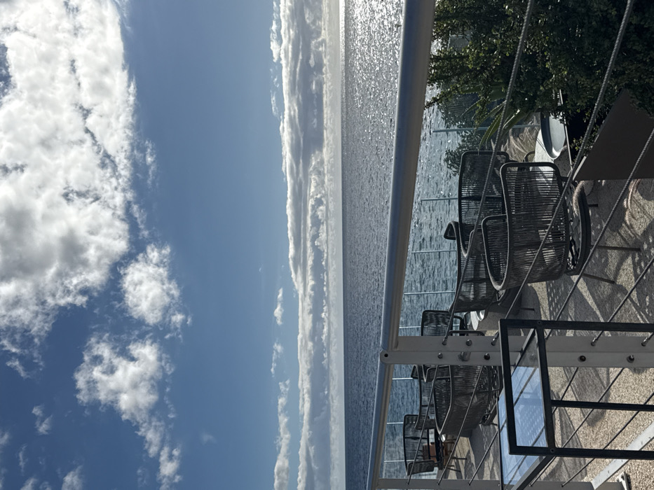 The image depicts an outdoor seaside seating area in Cagliari, Italy, under a partially cloudy blue sky. The upper half of the frame shows a blue sky with scattered white cumulus clouds, and a horizontal band of denser clouds along the distant horizon. The lower half features the calm, dark blue sea, with direct sunlight creating a bright, shimmering path on the water's surface towards the right.
In the foreground, a prominent silver metal railing with multiple horizontal cables extends across the entire width of the image. Below the railing, an outdoor terrace is visible, furnished with several empty dark grey woven chairs arranged around small, light-colored circular tables. These chairs feature curved backs and are positioned on a light-toned, textured ground surface. A transparent glass barrier is present between the seating area and the sea, offering an unobstructed view. To the lower left, a black metal-framed side table with a glass top is visible. Green foliage, consistent with low bushes or shrubs, is visible on the right side of the terrace, adjacent to the railing. No people, active subjects, or specific interactions are present in the scene.