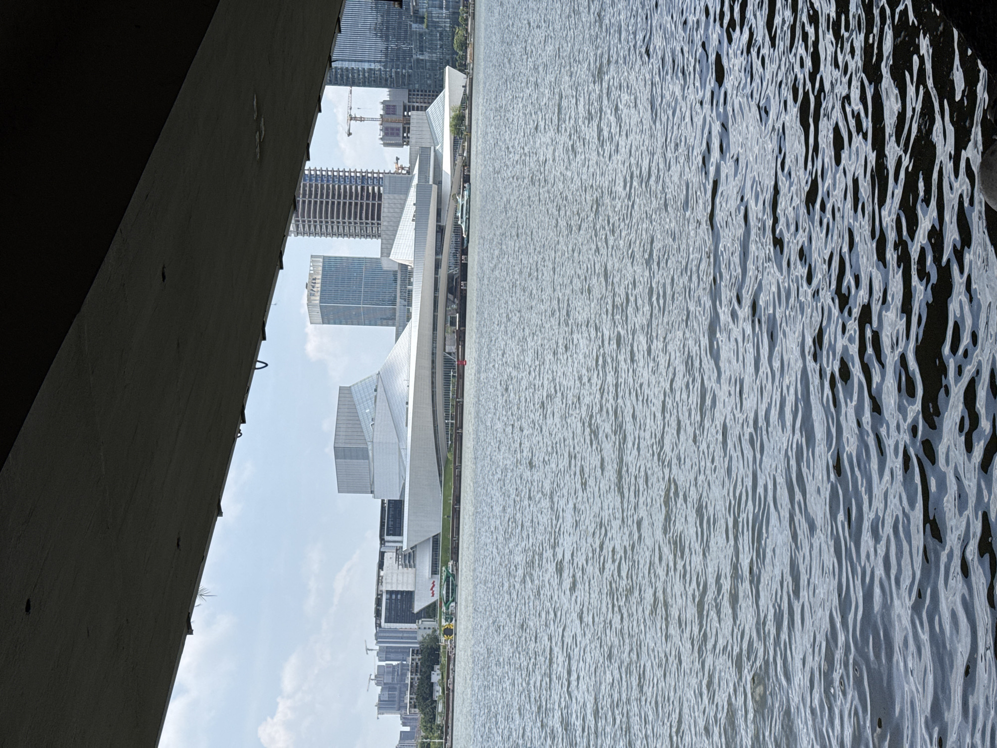 The image, taken from under a concrete structure, possibly a bridge, shows a body of water and a skyline with modern buildings. The water surface is rippled, reflecting light. Several high-rise buildings are visible, including one with a distinctive white, angular architectural design. Construction cranes are present, indicating ongoing development. The location is likely in the Liwan District, China, given the urban landscape and architectural styles. There are no people visible in the image.