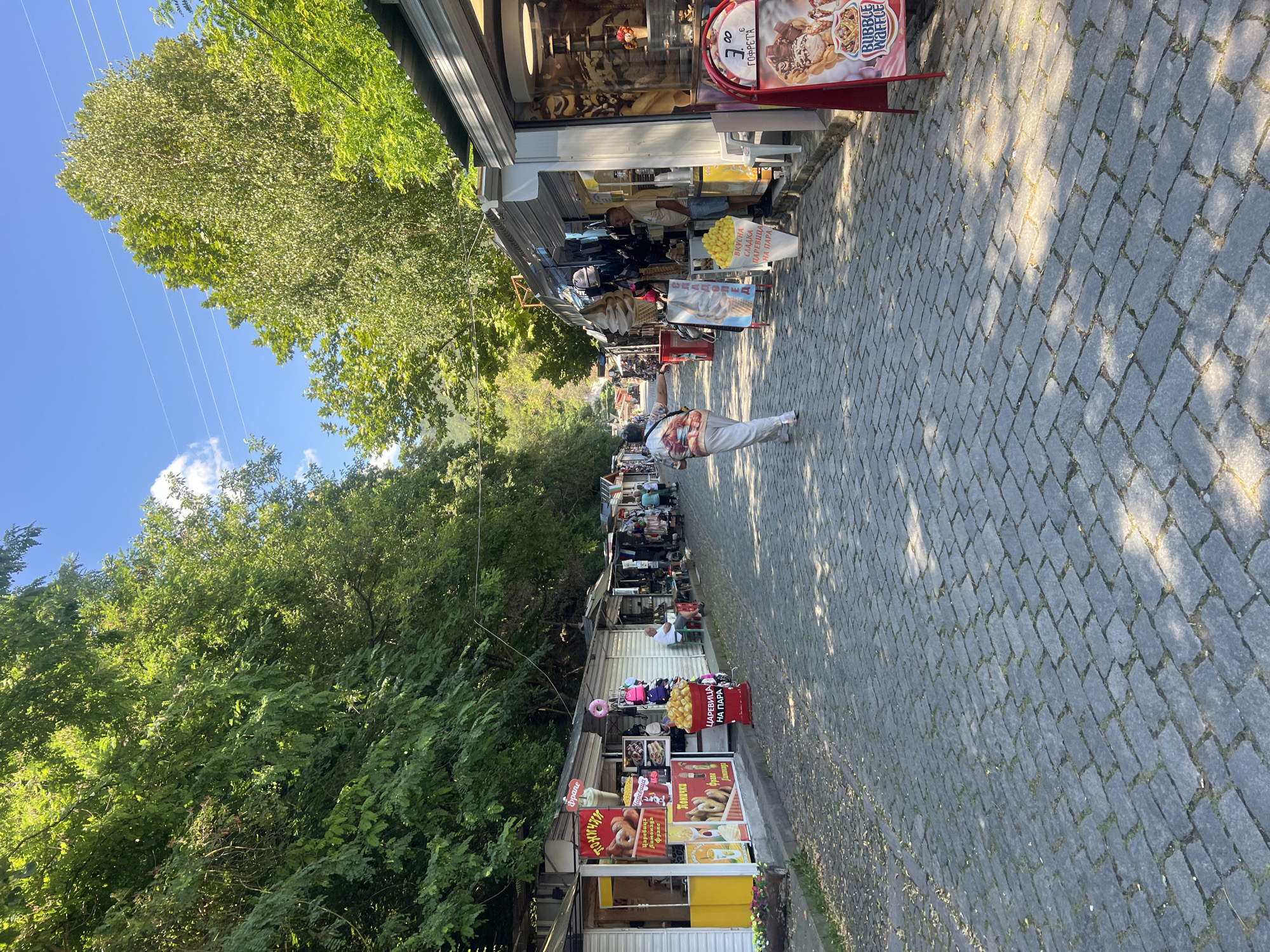 This is a medium-shot, eye-level, outdoor photograph of a cobblestone street lined with small shops. The shops appear to be tourist-oriented, selling items such as doughnuts, ice cream, and clothing. The street is shaded by mature trees on both sides, and the sky is blue with a few scattered clouds. A single person, wearing light-colored pants and a patterned shirt, is walking away from the camera down the center of the street. Several other people are visible in the background, browsing the shops or walking along the street. The location is identified as Bachkovo, Bulgaria, likely near the Bachkovo Monastery, a popular tourist destination. The atmosphere is bright and sunny, suggesting a pleasant day for outdoor activities.