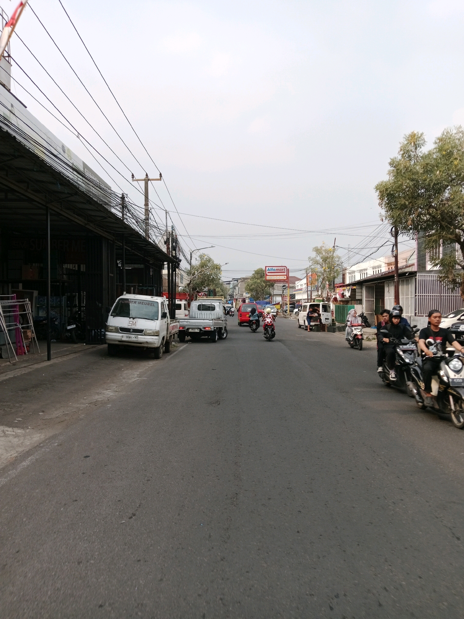 This image depicts a street scene in Cianjur, Indonesia. The road is paved and appears to be moderately busy with vehicular and pedestrian traffic. On the left side of the street, there are several open-fronted shops or workshops, one with the signage "SUMBER MEUBEL". These establishments have metal shelving units displaying various items. A white pickup truck is parked in front of one of these shops, with its license plate visible.
Further down the street, several motorcycles are in motion. Some are carrying multiple passengers. On the right side of the road, buildings line the street, including a structure with an Alfamart convenience store sign. Trees are also visible along the sides of the road. Overhead, a complex network of electrical wires is suspended between poles. The sky is overcast. The overall impression is that of a typical urban street in Indonesia during daytime.