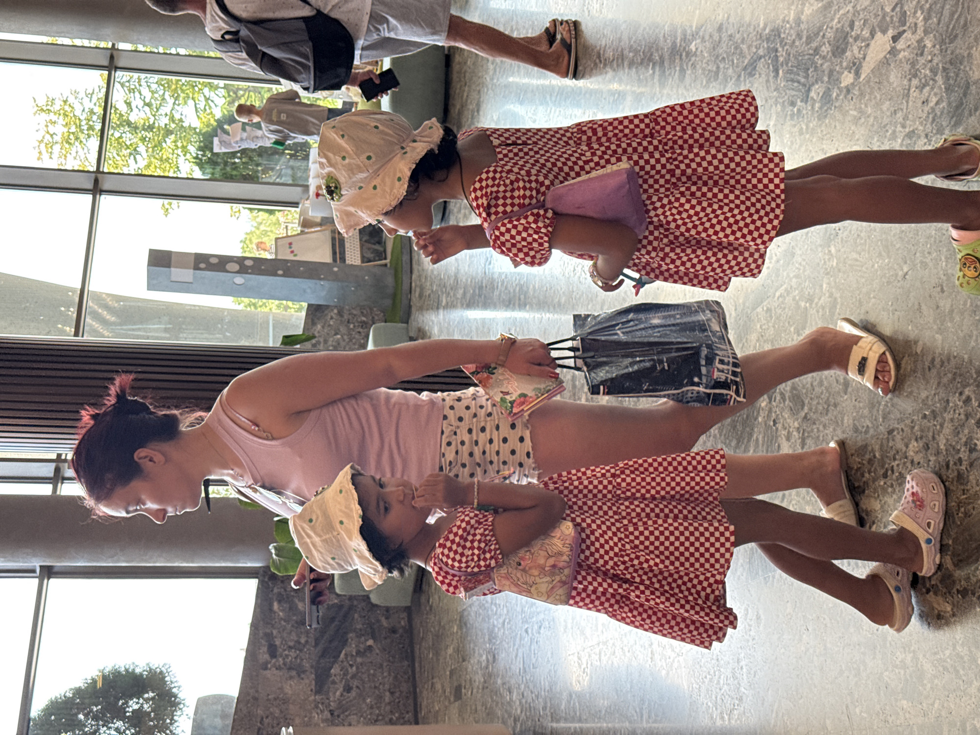 The image depicts a woman and two young girls inside a building with large windows and a marble floor. The woman, with dark hair pulled back, wears a light pink tank top and polka-dot shorts, and holds a phone in her left hand and a book or magazine in her right. She is standing behind the two girls. The girl on the left, wearing a red and white checkered dress and a white bucket hat with green polka dots, is facing left and appears to be biting her fingernail or licking her finger. She has a small purse with a unicorn print on her left shoulder. The girl on the right, also in a red and white checkered dress and a similar bucket hat, is facing left and also appears to be touching her mouth. She has a small purple purse on her left shoulder. Both girls are wearing sandals. In the background, through the windows, trees and greenery are visible. Other people are present in the background, including someone carrying a backpack and another individual with a backpack. The overall environment suggests a public indoor space, possibly a hotel lobby or a shopping center, located in the city of Holubynske rural hromada, Ukraine.