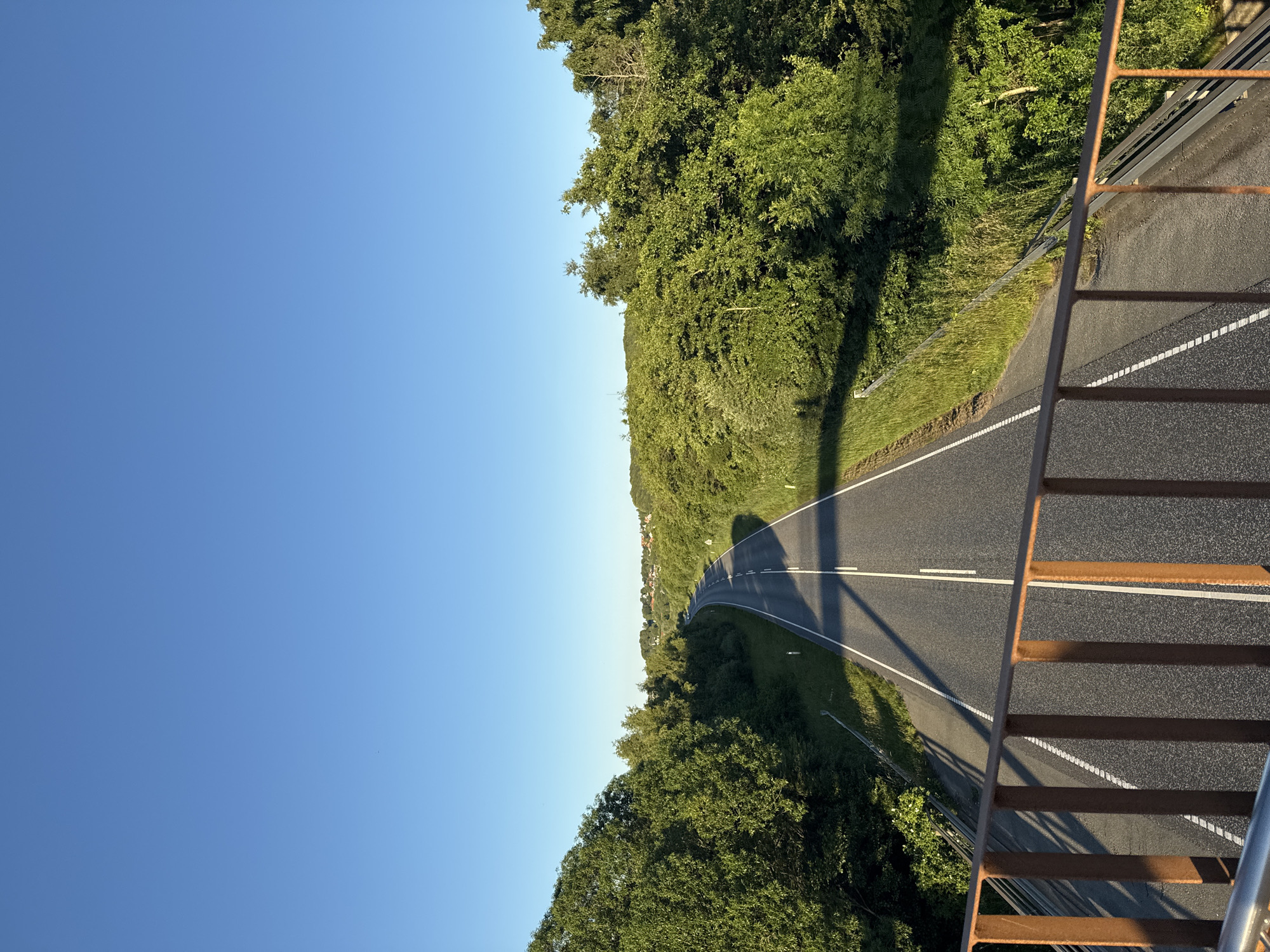The image depicts a clear, sunny day with a bright blue sky over a rural landscape in Vejle, Denmark. A grey asphalt road, marked with white lines, curves through lush green trees and vegetation. The road appears to be a multi-lane highway, with a median divider. In the foreground, a rusty brown metal railing runs along the edge of what appears to be a bridge or overpass, casting long shadows onto the road below. The surrounding area is densely wooded, with rolling hills visible in the distance, dotted with houses and buildings, suggesting a suburban or semi-rural setting near a town. There are no people or vehicles visible in the image. The overall scene is tranquil and natural.