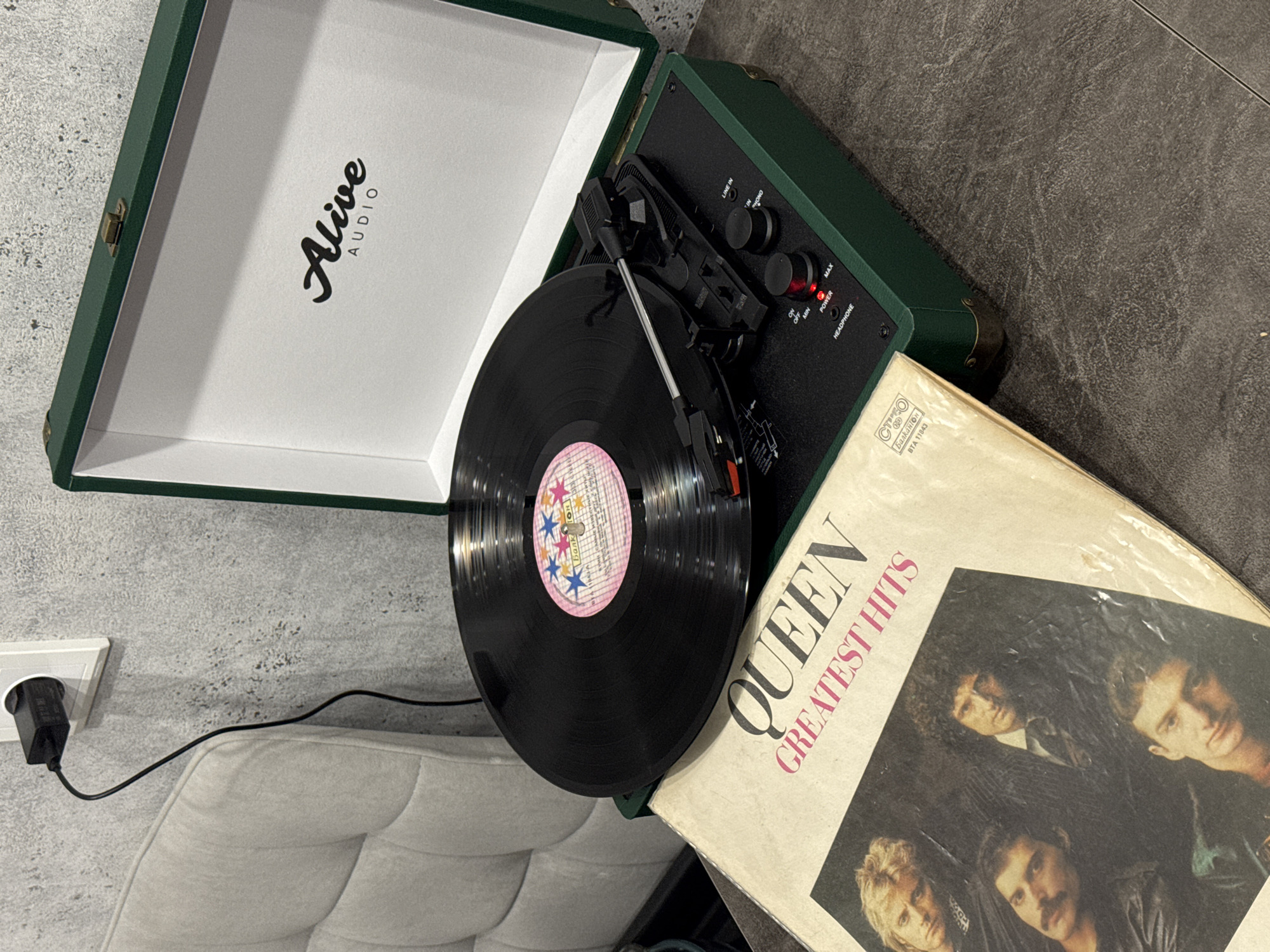 An overhead, angled shot captures a portable record player, a vinyl record, and an album cover in an indoor setting. The main subject is an open, dark green suitcase-style record player with "Alive AUDIO" prominently displayed in black script on the white interior of its lid. A black vinyl record rests on the turntable, featuring a pink and blue label with stars and the text "Балкантон" (Balkanton) and "BTA 11843", indicating a Bulgarian release.
To the right of the record player lies a record sleeve for "QUEEN GREATEST HITS," featuring a classic photo of the band members. Below the album title, the text "Балкантон" and "BTA 11843" are also visible, matching the record label. The record player's control panel, visible in the upper right, includes knobs and labels such as "LINE IN," "PHONO," "ON OFF MIN," "POWER" (with a red indicator light), "MAX," and "HEADPHONE."
In the foreground, part of a light-colored, upholstered chair or sofa is visible. A black power adapter is plugged into a white wall socket on a textured, grey wall, with its cord extending towards the record player. The floor beneath is a dark, textured surface. The scene suggests a relaxed atmosphere for listening to music.