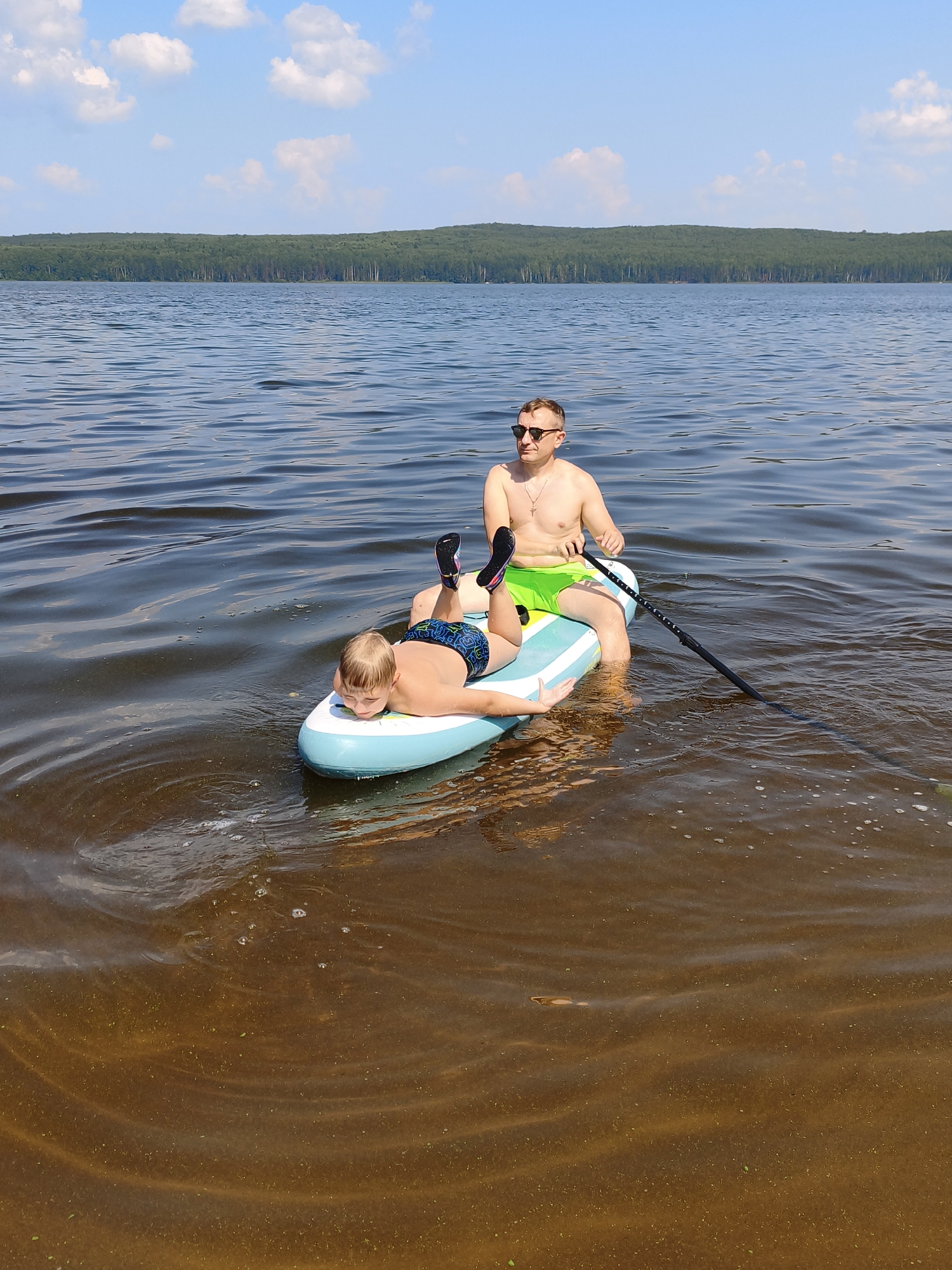 A man and a young boy are paddleboarding on a lake. The man, wearing sunglasses and green swim trunks, is seated on the paddleboard and holding a paddle. The boy, wearing blue swim trunks, is lying on his stomach on the front of the paddleboard. The water is calm with gentle ripples. In the background, a dense forest lines the shore of the lake under a bright blue sky with scattered white clouds. The scene appears to be set on a sunny day, likely in the afternoon, given the brightness of the light. The overall mood is relaxed and recreational. The location is Chaikovsky, Russia. There is no visible text in the image.