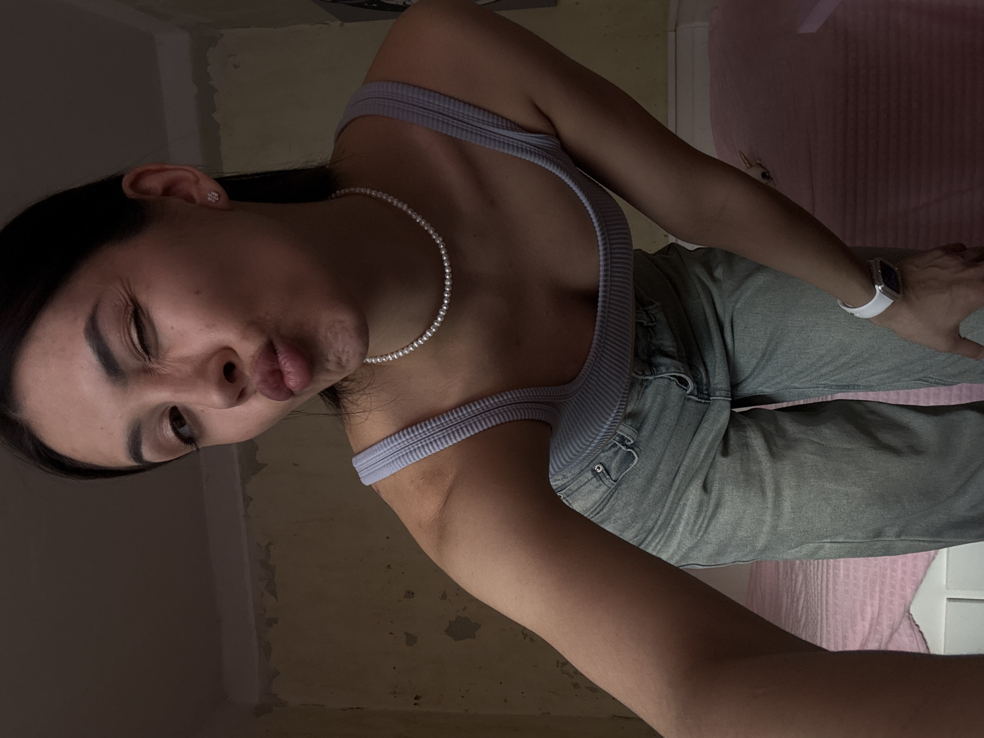 A young woman poses for a selfie in a dimly lit room with textured beige walls. She is wearing a light blue ribbed tank top and light wash jeans. A pearl necklace adorns her neck and a smartwatch is on her left wrist. She is making a pouting, winking expression. The location is likely indoors, but cannot be confirmed as Votkinsk, Russia without additional context.