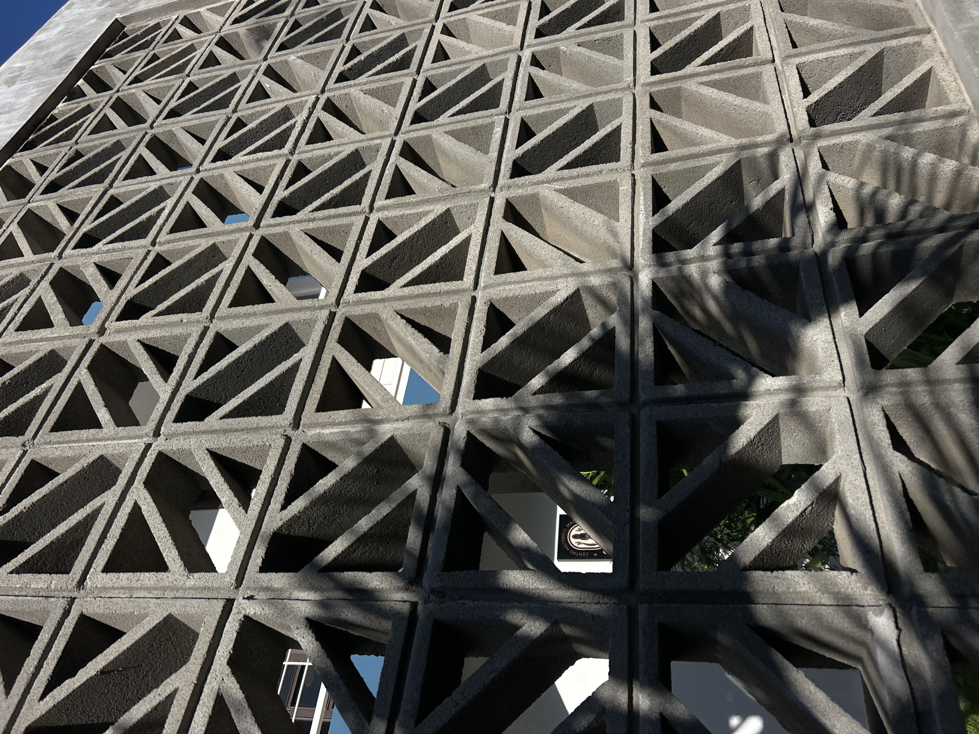 A closely framed image depicts a concrete breeze block wall in Mazatlán, Mexico, likely part of a building facade. The wall is composed of a repeating pattern of square modules, each containing four triangular openings oriented towards the center of the square. Light and shadow interplay across the wall's surface, casting distinct shadows from foliage outside the frame. The shadow patterns suggest the presence of palm trees or similar vegetation. In some openings, glimpses of the building's interior are visible, revealing white walls and a sign with an emblem. The photo focuses on the texture and design of the wall, capturing its geometric complexity and the interplay of light and shadow.