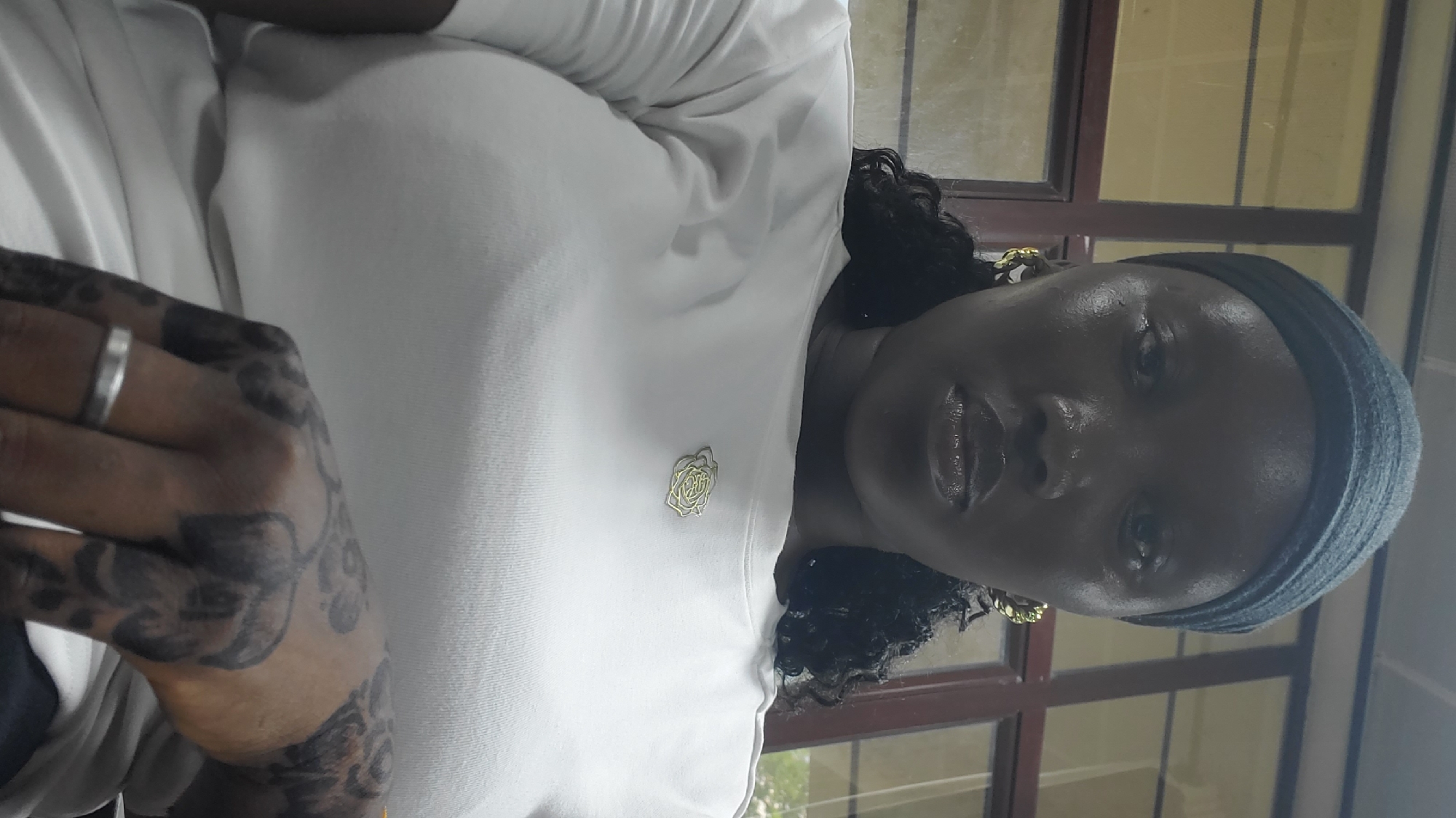 A medium shot captures a Black woman with dark skin, wearing a dark blue headscarf and a white t-shirt with a gold rose-shaped brooch. She is positioned in what appears to be an indoor setting, possibly an office or classroom, indicated by the presence of large windows with wooden frames in the background. The woman's face is directed towards the camera, with her lips slightly parted and her eyes looking straight ahead. She is wearing gold hoop earrings, and her hair is visible as dark curls cascading down. Her left hand, adorned with intricate dark henna tattoos and a silver ring, rests on her chest, obscuring a portion of her t-shirt. The lighting in the image is somewhat dim, casting subtle shadows on her face and highlighting the sheen on her lips. The overall impression is a candid portrait taken indoors.