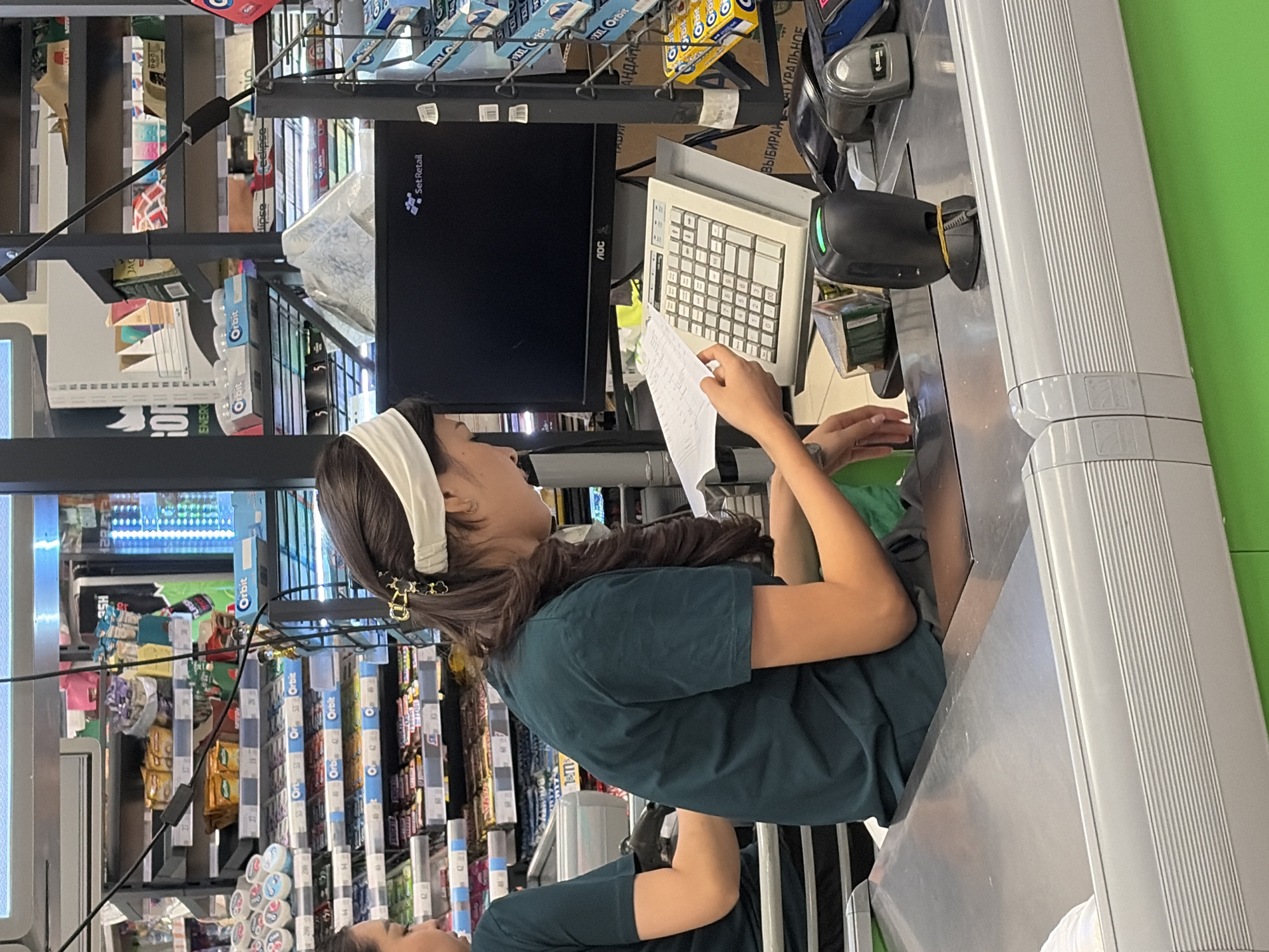 The image shows two young women in dark green t-shirts at a grocery store checkout counter. The woman on the right is wearing a white headband and has dark hair with a decorative clip. She is holding a piece of paper and looking down at it. In front of her is a cash register with a keyboard and a computer monitor displaying the "SetRetail" logo. To the right of the keyboard are two barcode scanners. The woman on the left is partially visible, with only her left shoulder and arm showing as she appears to be working at the checkout. Behind the women are shelves stocked with various products, including candy, gum, and packaged goods. The checkout counter is made of stainless steel, and the floor is bright green. The lighting suggests an indoor environment, likely a supermarket.