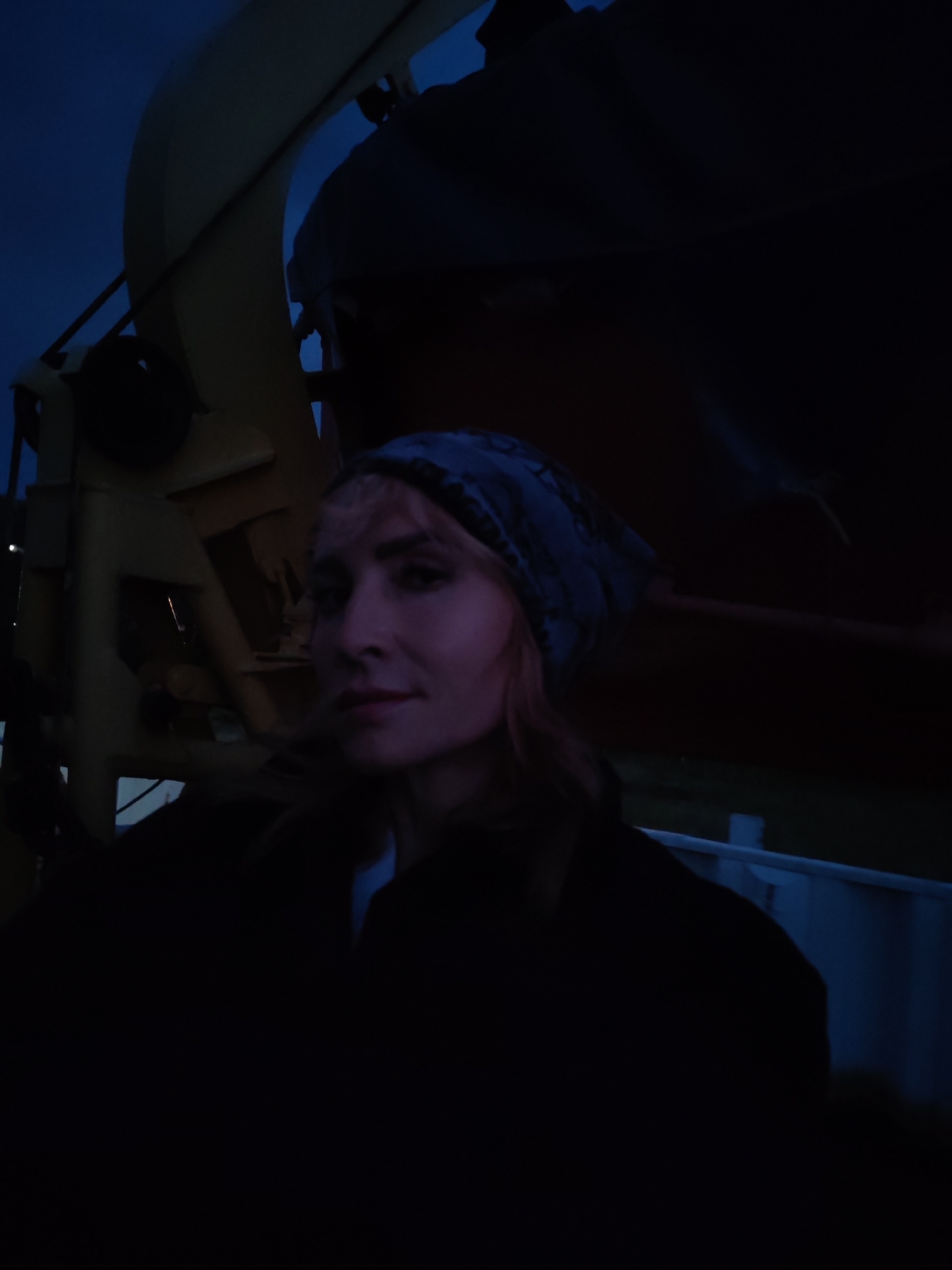 An outdoor, waist-up, medium shot captures a woman wearing a patterned bandana on her head. Her hair is light brown and appears to extend past her shoulders. She is looking directly at the camera, with her head tilted slightly to her right. Her facial features are discernible despite the low light conditions. She is wearing dark clothing. Behind her, on the left side of the frame, a large, yellow, industrial structure with dark gray accents is visible. The background consists of a dark blue sky and a darker, indistinct mass that could be part of the same structure or another object. The overall lighting is dim, suggesting it is either dusk or nighttime. The image is shot from a slightly upward angle.