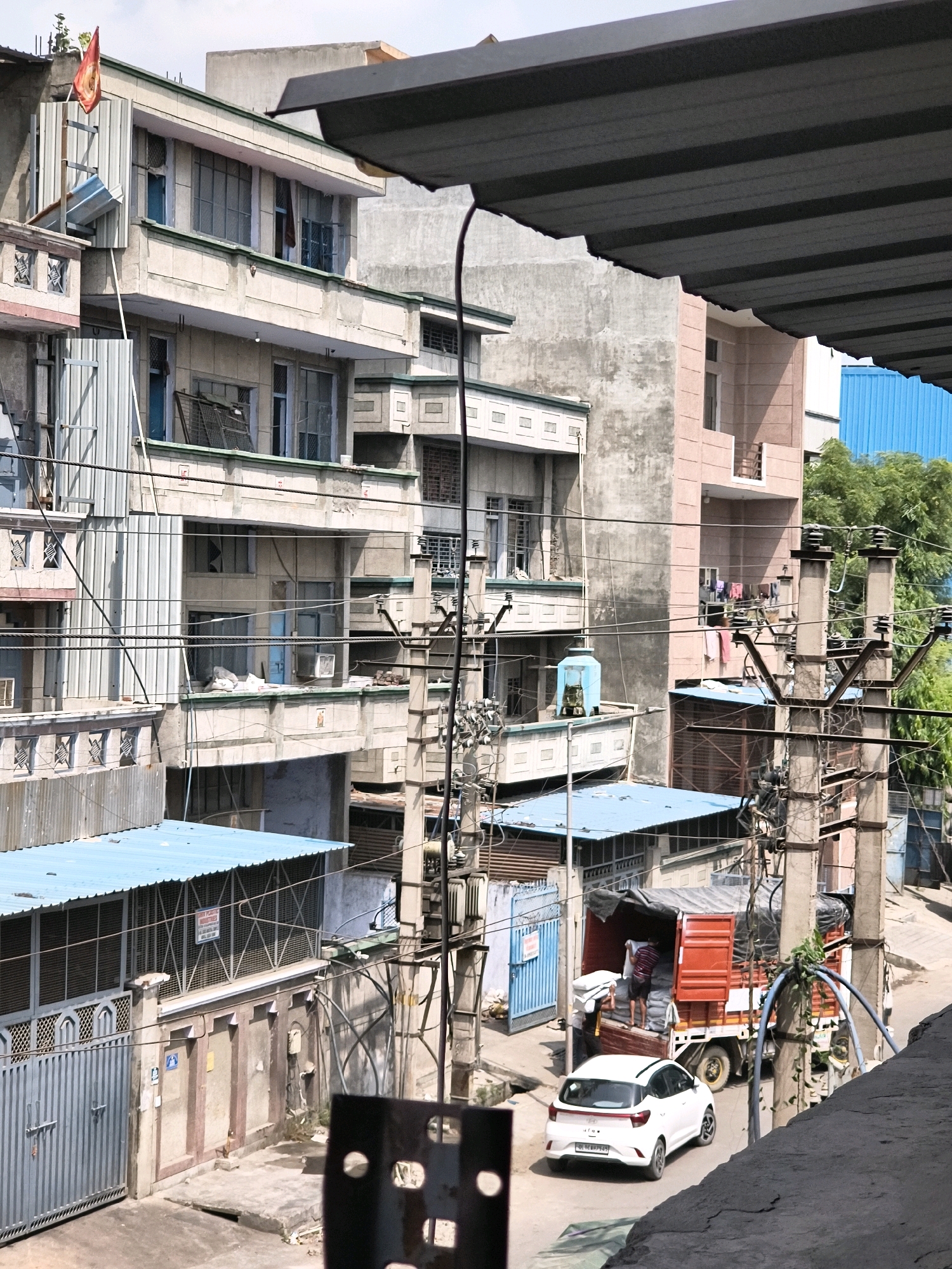 The image depicts a street scene in a dense urban environment, likely in Delhi, India, as suggested by the architecture, signage, and the presence of a red delivery truck with a license plate that appears to be in the Indian format. The scene is characterized by multi-story buildings constructed with concrete, featuring balconies, numerous windows, and external electrical wiring and utility poles. A red truck, partially loaded with what appear to be large bags of material, is parked on the street. A white hatchback car is also visible. Several individuals are present, including at least two men near the truck, one of whom is actively engaged in loading it. Laundry is hanging on a line on a balcony of one of the buildings. The overall impression is one of a busy, somewhat cluttered urban area with ongoing commercial activity.