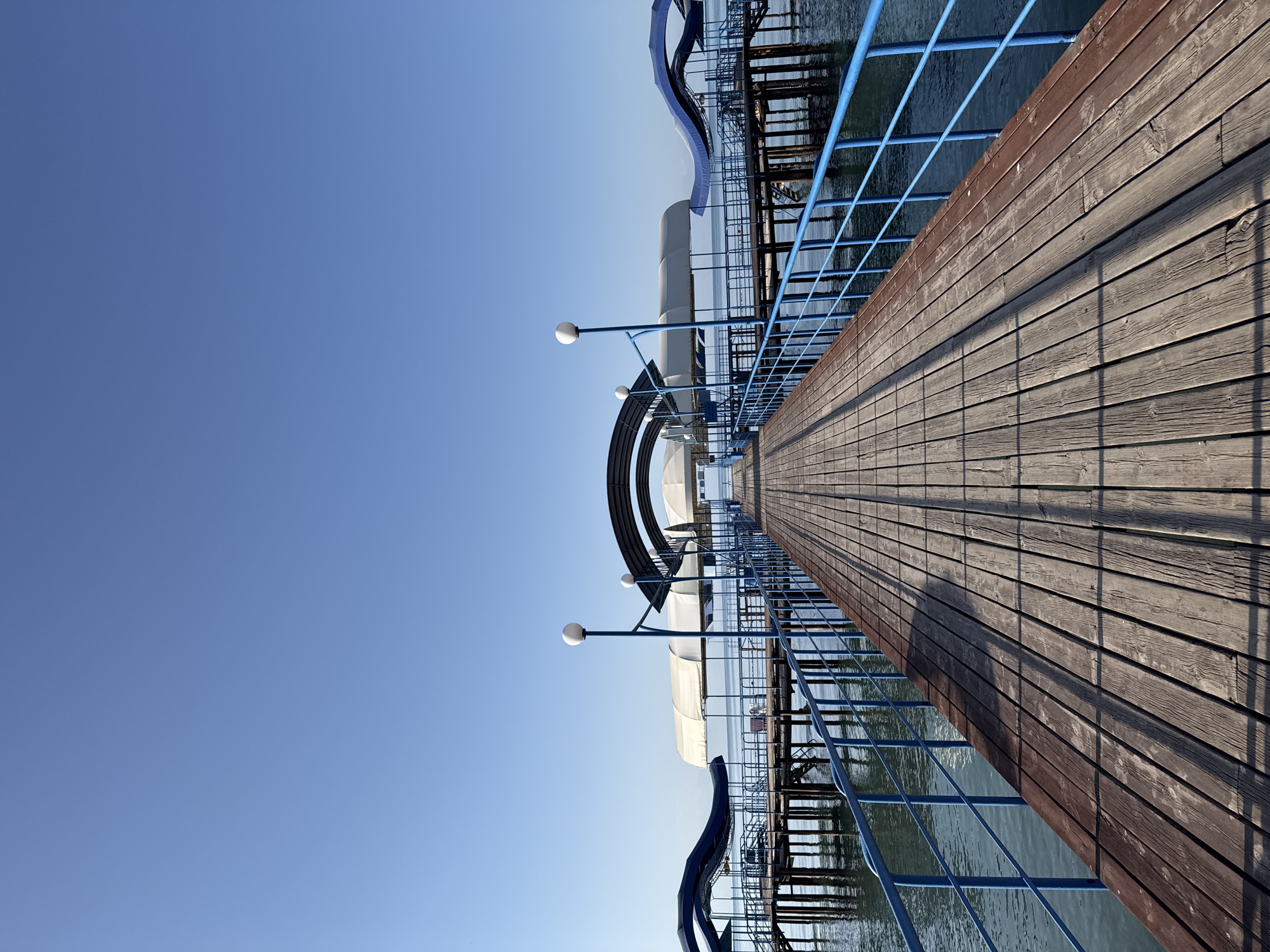 This is a low-angle, wide shot of a wooden pier extending into a calm body of water under a clear blue sky. The pier is made of weathered wooden planks and has a bright blue railing running along both sides. The pier has covered sections with white and blue canopies. The water reflects the blue of the sky and the structure of the pier. The scene appears to be a public space, possibly a resort or recreational area, in Sary-Oy, Kyrgyzstan. There are no people visible in the image, nor any specific actions or interactions taking place.