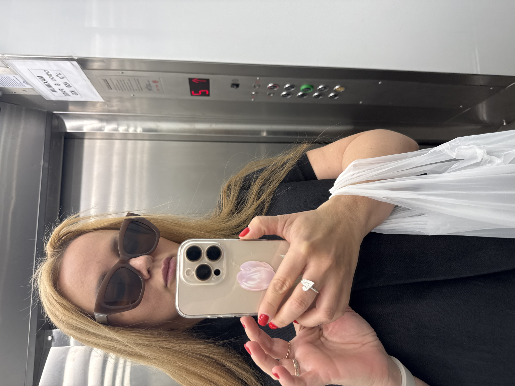 The image depicts a selfie taken by a blonde woman wearing brown sunglasses in an elevator. She's holding her smartphone, which has a light-colored case and is adorned with a pink floral design. She is wearing a black shirt. The woman is wearing red nail polish, and a ring. Behind her, the elevator's control panel is visible. A sign is posted above the control panel. The number "5" is illuminated in red on the display. The elevator appears to be located in Tel-Aviv, Israel.