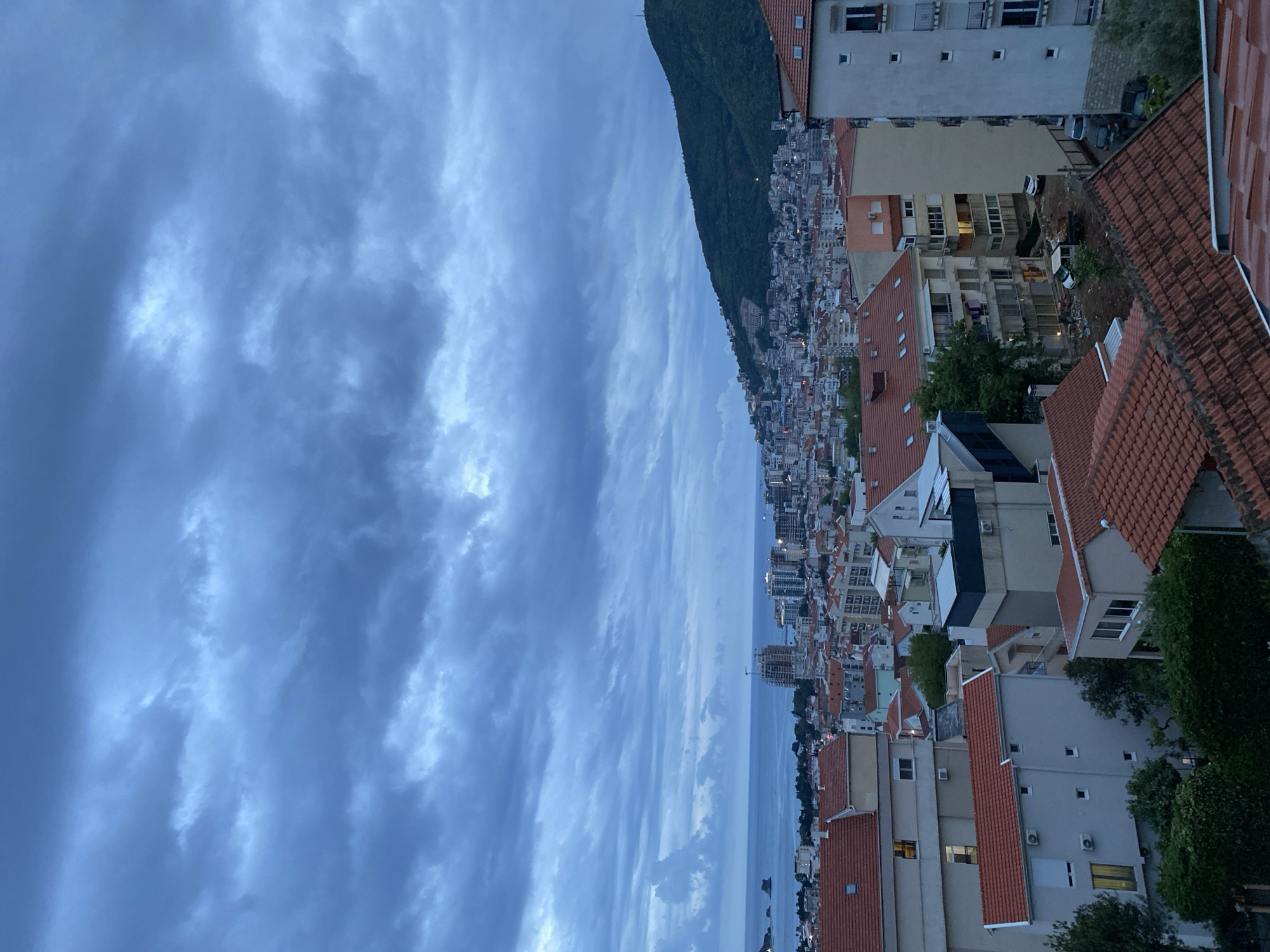 This wide-angle image captures a sprawling coastal city, identified as Budva, Montenegro, nestled between a majestic mountain and the Adriatic Sea under an overcast sky. The scene depicts dusk or early evening, as artificial lights are beginning to illuminate windows and streets throughout the urban landscape.
In the foreground, a dense array of residential and commercial buildings dominates the lower right portion of the frame. Many structures feature distinct red-tiled roofs, characteristic of Mediterranean architecture, interspersed with lighter-colored facades. Details like air conditioning units, windows (some with lights on), and glimpses of parked cars in alleys are visible. Green foliage, including trees and bushes, punctuates the urban sprawl.
To the left, a large, dark green mountain, thickly covered in trees, rises dramatically, forming a rugged backdrop to the city. Its slopes are dotted with more buildings, indicating the city extends up the mountainside.
Beyond the immediate urban area, the cityscape stretches extensively along the coastline, eventually meeting the calm, expansive sea on the right. In the distance over the water, faint outlines of smaller landmasses or islands are discernible, along with a construction crane, suggesting ongoing development.
The sky is a prominent feature, filled with layers of blue-grey clouds that create a diffused, soft light across the entire scene. There are no visible people or discernible text, and the overall mood is tranquil and atmospheric, capturing the city transitioning from day to night.