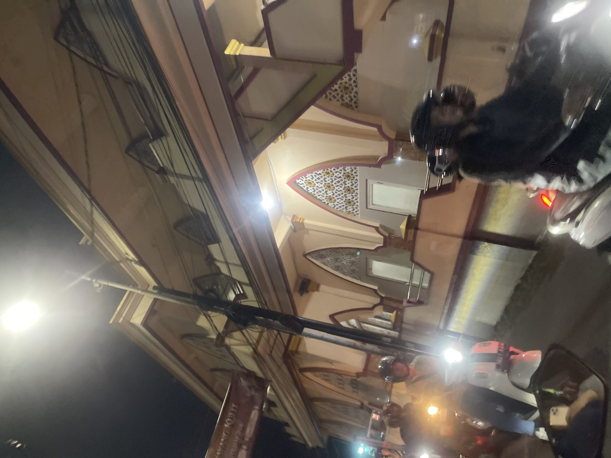 This image captures a street scene at night in Sawotratap, Indonesia. The primary focus is on a building with distinct architectural elements, featuring arched openings decorated with intricate, geometric patterns in yellow and white against a muted background. The building's facade is adorned with maroon and cream colored trim. Above the building, a complex network of electrical wires is visible, strung between poles.
In the foreground, several individuals on motorcycles are present, suggesting a street or thoroughfare. One person, wearing a helmet, is seated on a motorcycle on the right side of the frame, partially obscured by glare. Another motorcycle, with a person wearing a helmet and dark clothing, is also visible. A bright, white light from a streetlamp illuminates the left side of the image, casting a strong glow and contributing to the nighttime atmosphere. The overall impression is of a bustling urban environment at night, with the architectural beauty of the building contrasting with the utilitarian network of wires and the presence of motorbikes. No specific event or emotion is clearly conveyed, but the nighttime setting and the presence of vehicles suggest general urban activity. A portion of a sign with text is visible in the lower left, but the text is illegible.