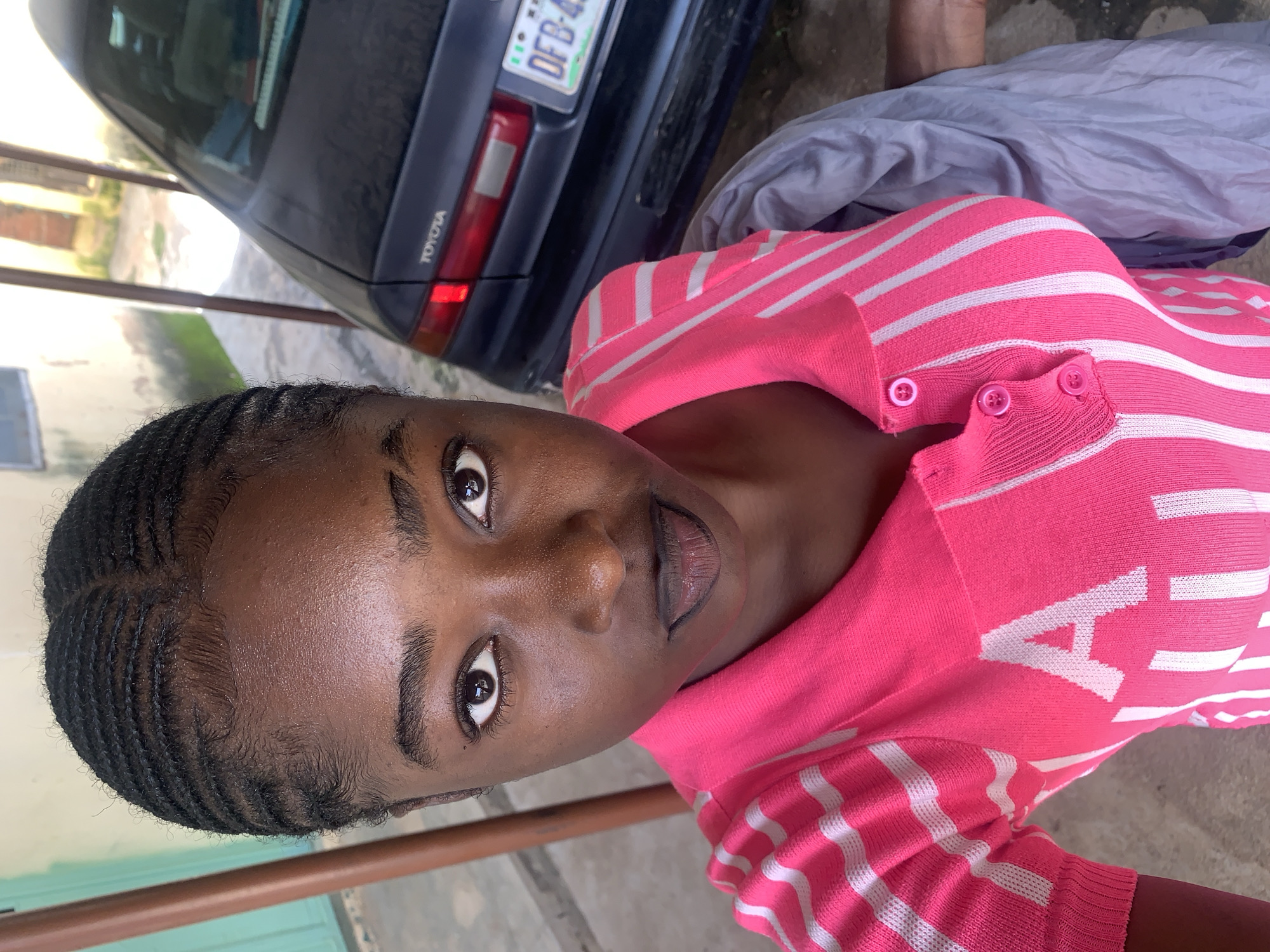 A young woman of African descent is captured in a close-up, waist-up portrait outdoors, likely in Rafin Par, Nigeria. Her dark hair is styled in neat cornrows, and she has dark eyes and dark lips. She is wearing a vibrant pink polo shirt with white stripes and buttons. The background shows a dark-colored Toyota sedan with a license plate that reads "DFB-455," partially visible. The environment appears to be an open-air area, possibly a driveway or courtyard, with a building structure and a pillar visible on the left side. The lighting suggests it is daytime.