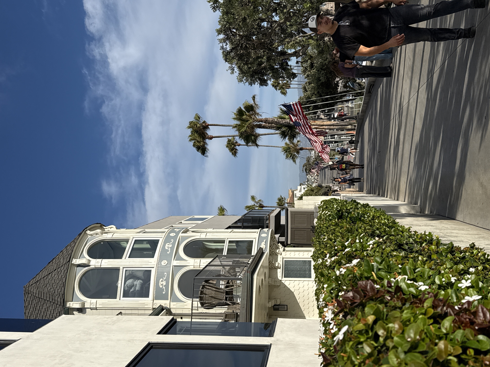 This image depicts a sunny day scene on a street in Manhattan Beach, United States of America, likely near the ocean given the palm trees and clear blue sky. In the foreground on the left, a white building with a dark, mansard-style roof and ornate white trim is visible, featuring arched windows and a balcony. Adjacent to it, a lush green hedge with white flowers lines a concrete pathway.
The pathway extends into the background, where several people are walking. Palm trees line the right side of the pathway, with a row of American flags displayed along the street. A man wearing a black t-shirt, jeans, and a baseball cap is walking towards the right of the frame in the mid-ground. Other individuals and a dog can be seen further down the pathway. The overall atmosphere is relaxed and casual, suggesting a public area such as a boardwalk or a beachside street.