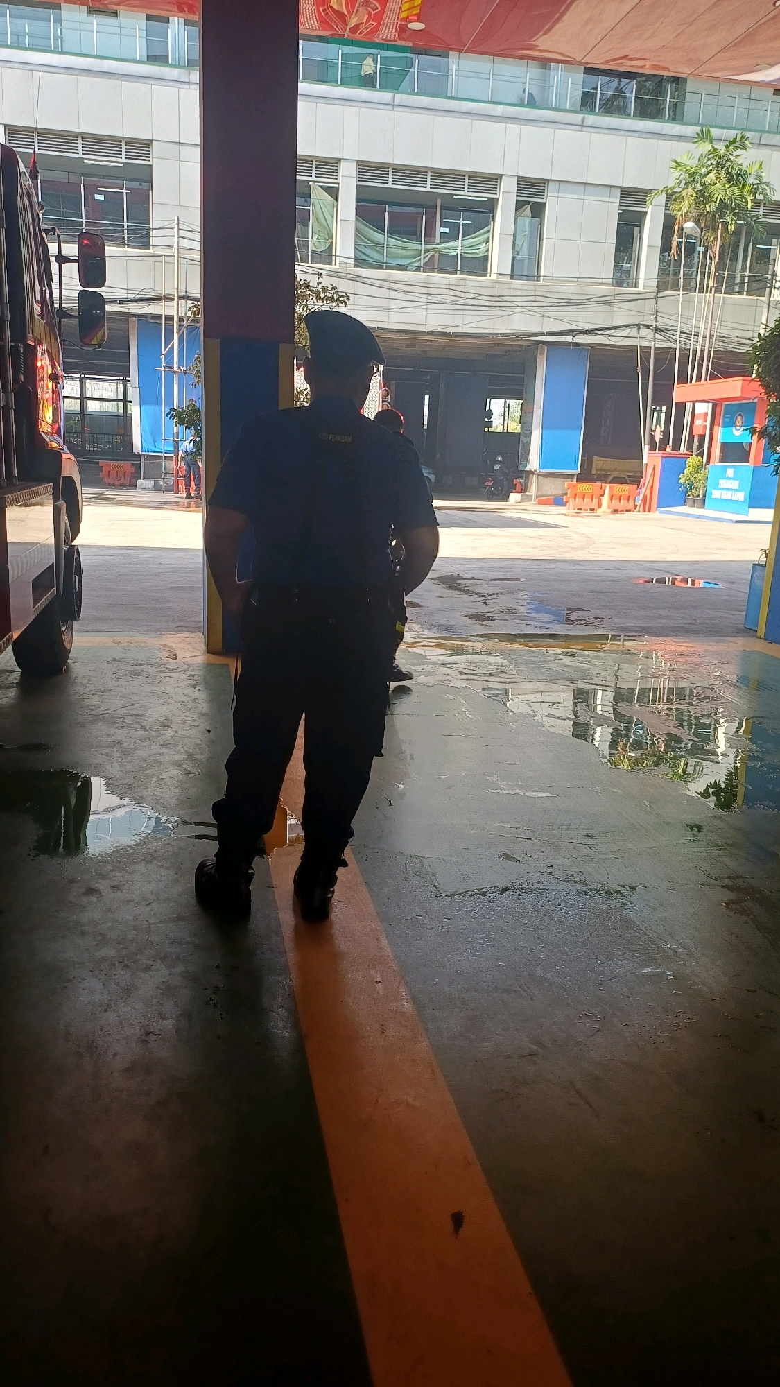 The image shows a male individual wearing a dark blue uniform and a beret standing in an outdoor, covered area. The uniform has text on the back that appears to read "DAMKAR" and possibly "PEMADAM", indicating he is likely a firefighter. He is facing away from the camera, with his hands in his pockets. The ground is concrete and wet, with visible puddles reflecting the surroundings. In the background, a multi-story building with many windows is visible, along with some palm trees. A red fire truck is partially visible on the left side of the frame. The overall setting suggests a fire station or a similar emergency service facility. This scene is consistent with locations in South Jakarta, Indonesia, where such facilities are common.