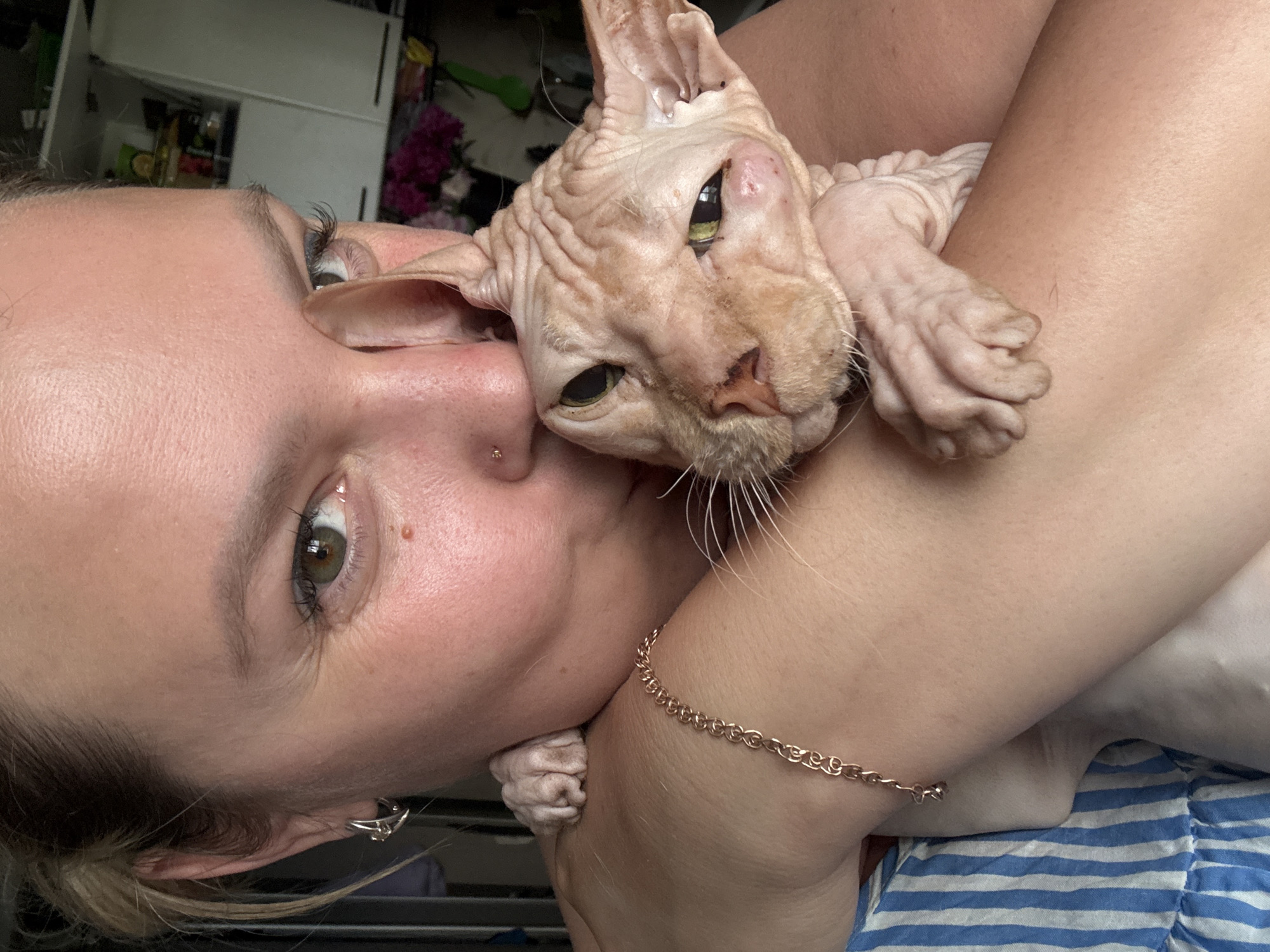 A woman with blonde hair pulled back is holding a hairless cat, likely a Sphynx breed, close to her face. The cat has greenish eyes and wrinkled skin. The woman has green eyes and is wearing a silver hoop earring in her left ear and a nose stud. A thin gold chain bracelet with a small charm is visible on her left arm. She is wearing a blue and white striped garment. The background is out of focus but appears to be an indoor setting with shelving units and some flowers. The image is a close-up, chest-up shot. There is no information provided about the location within Марковский, Russia.