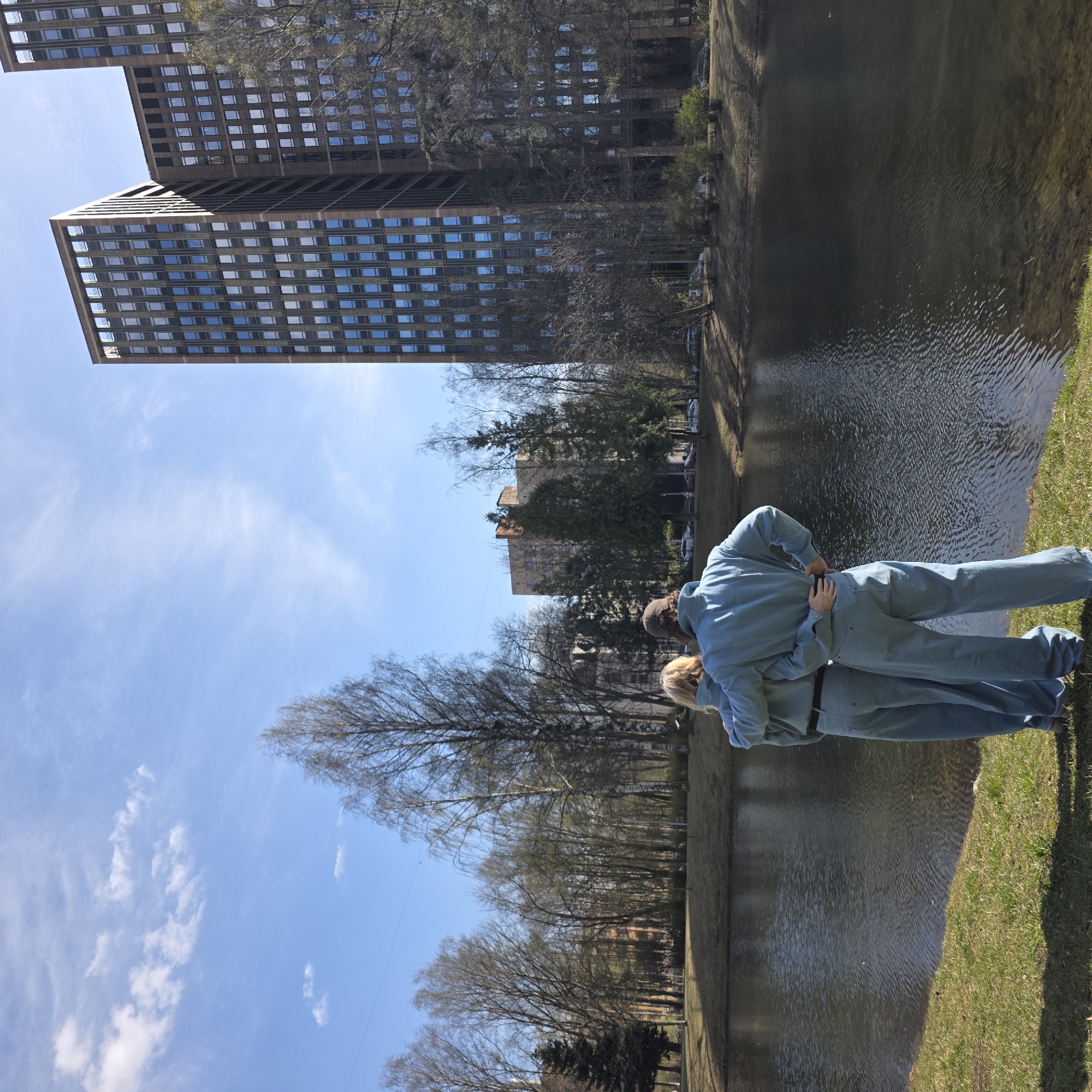 This image, captured in Saint Petersburg, Russia, depicts a bright urban landscape by a body of water on a clear day. Two individuals are prominently featured in the foreground, seen from behind, standing closely together by the water's edge. The taller person, with dark hair, is wearing a light blue jumpsuit or overalls and has their hands on the waist/back of the shorter, blonde-haired individual, who is dressed in a similar light blue outfit. Their posture suggests an intimate moment, possibly an embrace or shared contemplation of the view.
The setting includes a calm body of water, likely a pond or canal, that stretches across the middle ground. Its surface shows subtle ripples and reflects the bright blue sky. A narrow strip of green grass forms the bank where the individuals are standing. In the background, two tall, modern buildings with numerous rectangular windows dominate the skyline, their dark facades contrasting with the light. Several trees, mostly bare of leaves, indicating early spring or late autumn, are scattered along the banks and in the distance. The sky is a vibrant blue with sparse, wispy white clouds, suggesting sunny and pleasant weather. No discernible text is visible.