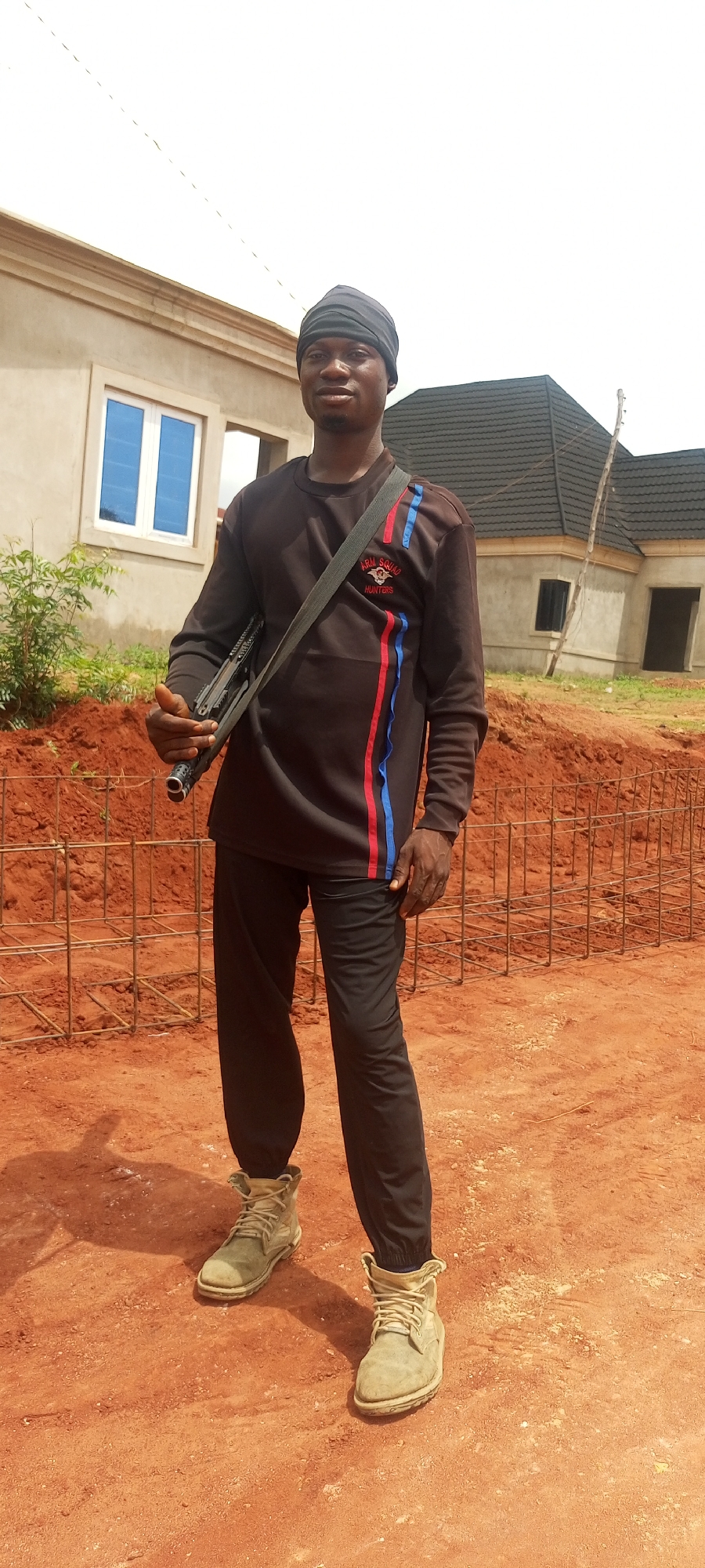 A young, dark-skinned man stands outdoors on reddish-brown bare earth, likely a construction site, under bright daylight. He wears a black head covering, a dark long-sleeved shirt with distinct red and blue vertical stripes on the right side, black pants, and light-colored, sturdy work boots. On the left chest of his shirt, a logo displays the text "ARM SQUAD HUNTERS." A firearm, appearing to be a shotgun, is slung across his body, held by his right hand. He looks directly at the camera with a neutral expression.
In the background, partially constructed buildings are visible. To his left, a light-colored building with blue-tinted windows. To his right, another building features a dark tiled roof. Metal rebar cages, commonly used in construction, are scattered on the ground. The scene depicts an area under development in Idah, Nigeria, characterized by dry, red soil and clear, sunny weather.