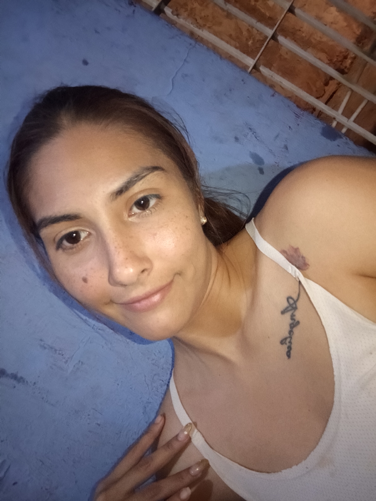 A woman is lying on a light blue surface, possibly a bed or similar. She has light brown hair pulled back, brown eyes, and freckles on her face. She is wearing a white tank top. A tattoo is visible on her chest, and a purplish mark is on her upper chest near her shoulder. Her right hand is resting near the left side of the image. The upper right corner shows a brick structure. The location is stated to be Juan Griego, Venezuela.
