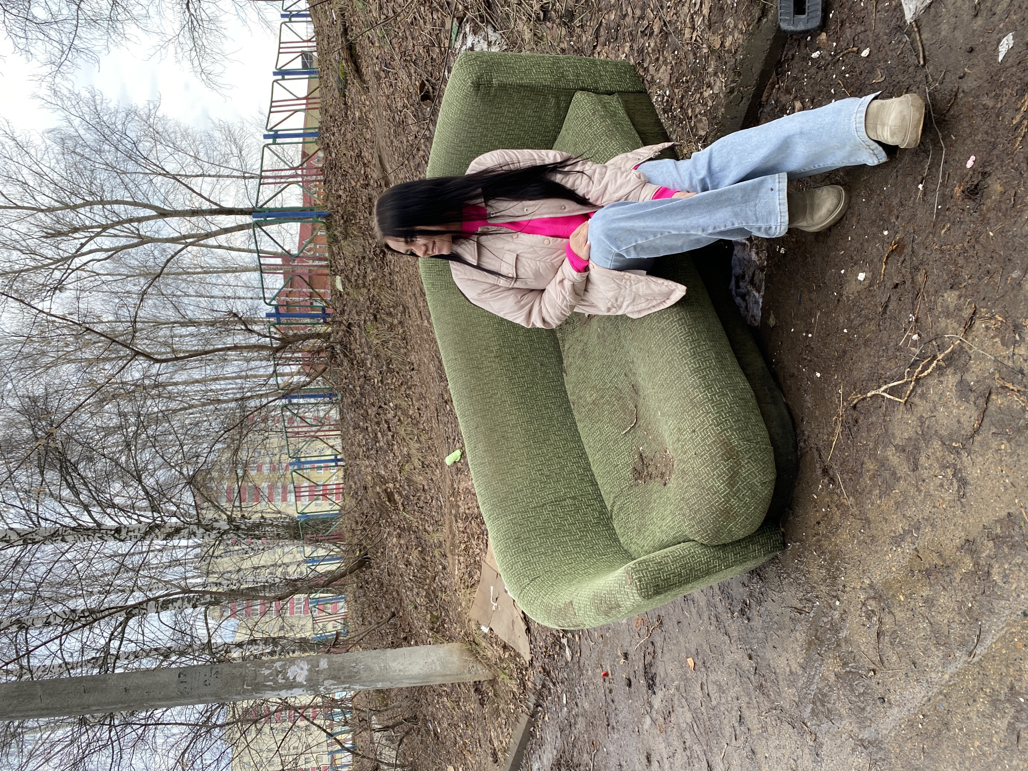A young woman is sitting on a green couch in what appears to be a neglected urban environment. She has long, dark hair and is wearing a pink sweater under a light-colored jacket, light blue jeans, and beige shoes. The couch is placed on a patch of dirt and decaying leaves, suggesting it was abandoned outdoors. In the background, bare trees and a building with colorful accents typical of playground or recreational facilities are visible. The scene suggests urban decay. The location is Chaikovsky, Russia.