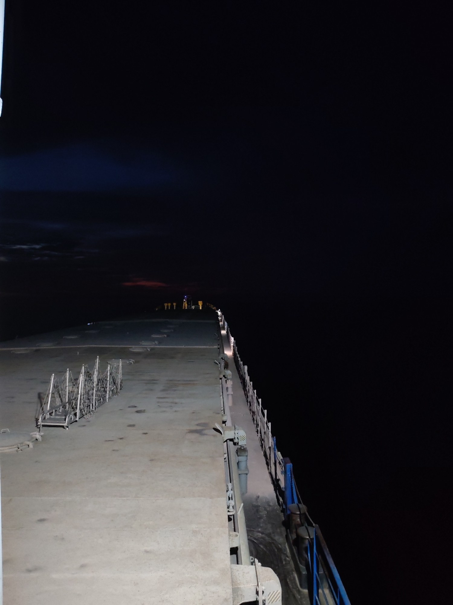 The image depicts a view from the deck of a vessel at night. A concrete deck stretches towards the horizon, with a metal railing running along the right side. In the distance, several yellow lights are visible, along with a blue light. The sea is dark, and the sky is black with a hint of a reddish cloud formation. There is no evidence of people or any activity. The location is identified as Kvartsitny, Russia, suggesting this is a port or harbor scene.