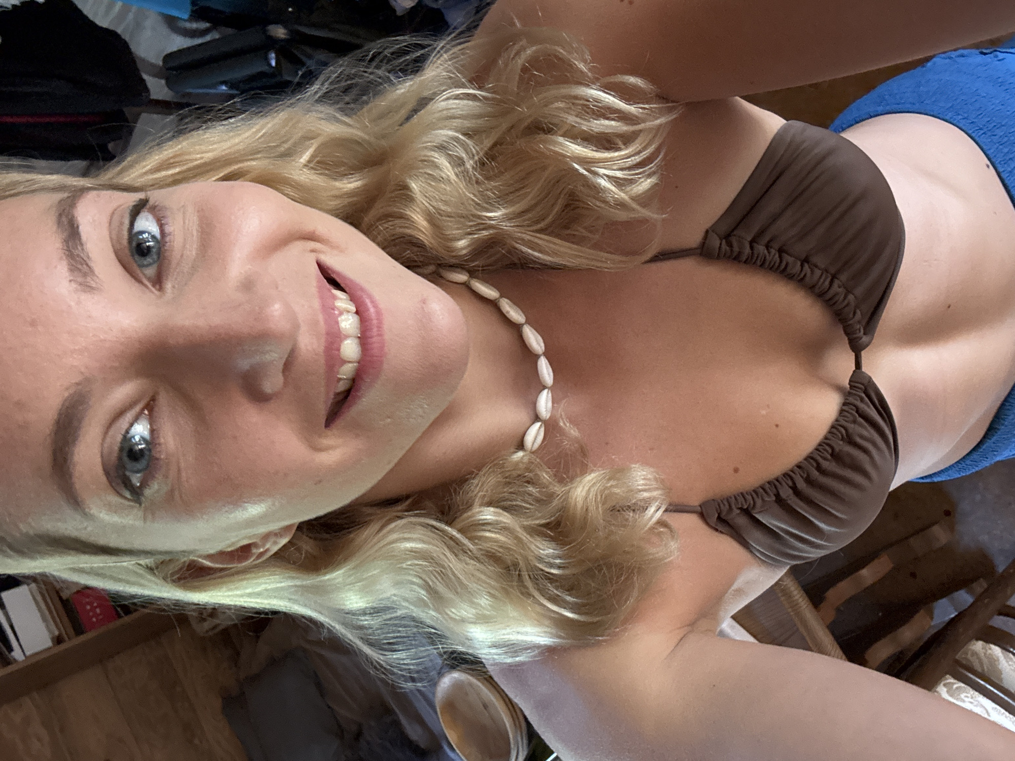 A woman with blonde hair, wearing a brown bikini top and a shell necklace, smiles at the camera. She also wears a blue garment around her waist. The location appears to be indoors.