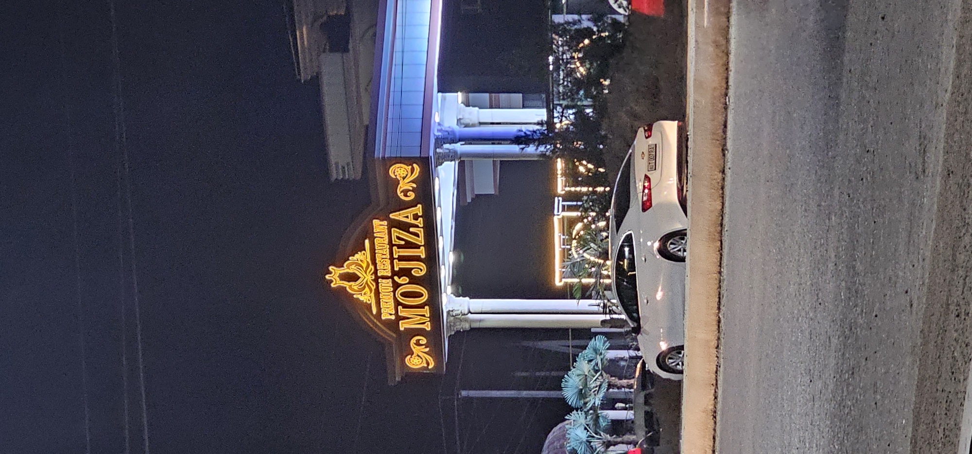 This is a nighttime, eye-level, outdoor, full-shot photograph of the entrance to a restaurant named "MO'JIZA" in Bukhara, Uzbekistan. The restaurant has a large, illuminated sign with the name "MO'JIZA" in prominent yellow lettering, with "PREMIUM RESTAURANT" written above it in smaller text. The sign is adorned with a decorative crown-like emblem at the top. The entrance features decorative pillars and is illuminated by blue and yellow lights. In front of the entrance, parked on the side of a road, is a white sedan. The foreground shows a paved road and a curb. The background is dark, indicating nighttime, with some distant lights and foliage visible. The image captures a slice of urban infrastructure and commercial establishments at night.