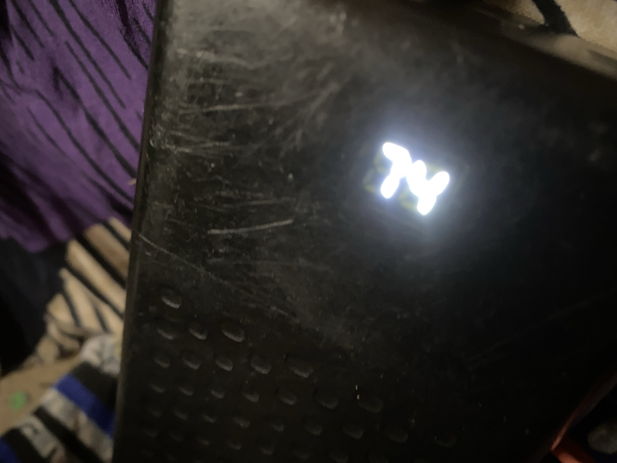 The image shows a close-up of a black textured surface with a lit symbol in white. The symbol appears to be abstract, possibly a stylized "N" or a pair of interlocking shapes. The surface is scratched and worn, suggesting it is an object that has seen use. In the background, there are out-of-focus patterns of fabric in purple and stripes of black, white, and blue, indicating the object is placed on or near soft furnishings. The lighting is dim, with the bright symbol being the primary light source in the immediate area. There are no people or explicit environmental details that can pinpoint a specific location in Jauro Yinu, Nigeria. The image is a detail shot of an object's surface and its illuminated indicator.