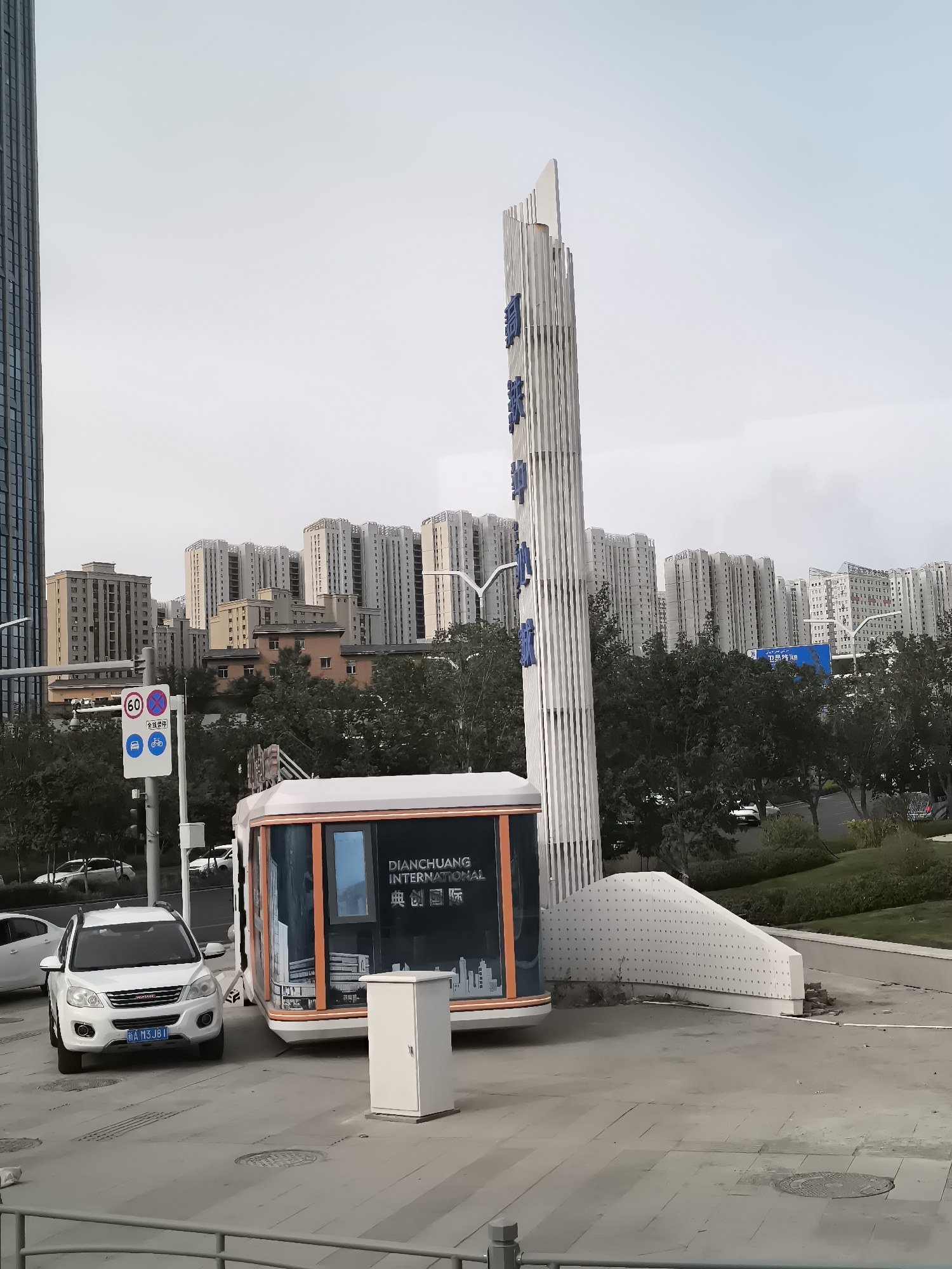 This image depicts a street scene in Ürümqi, China. In the foreground, a white SUV with a blue license plate is parked. To its right, a white kiosk-like structure with orange trim is situated. Behind these vehicles and structures, a modern sculptural monument with vertical slats and blue lettering stands tall. In the background, a dense array of multi-story residential buildings with light-colored facades dominates the skyline under an overcast sky. Trees and greenery are visible between the buildings and in a landscaped area. A traffic sign indicating a speed limit of 60 and other traffic regulations is also present. The overall environment suggests an urban setting with ongoing development or a modern residential area.