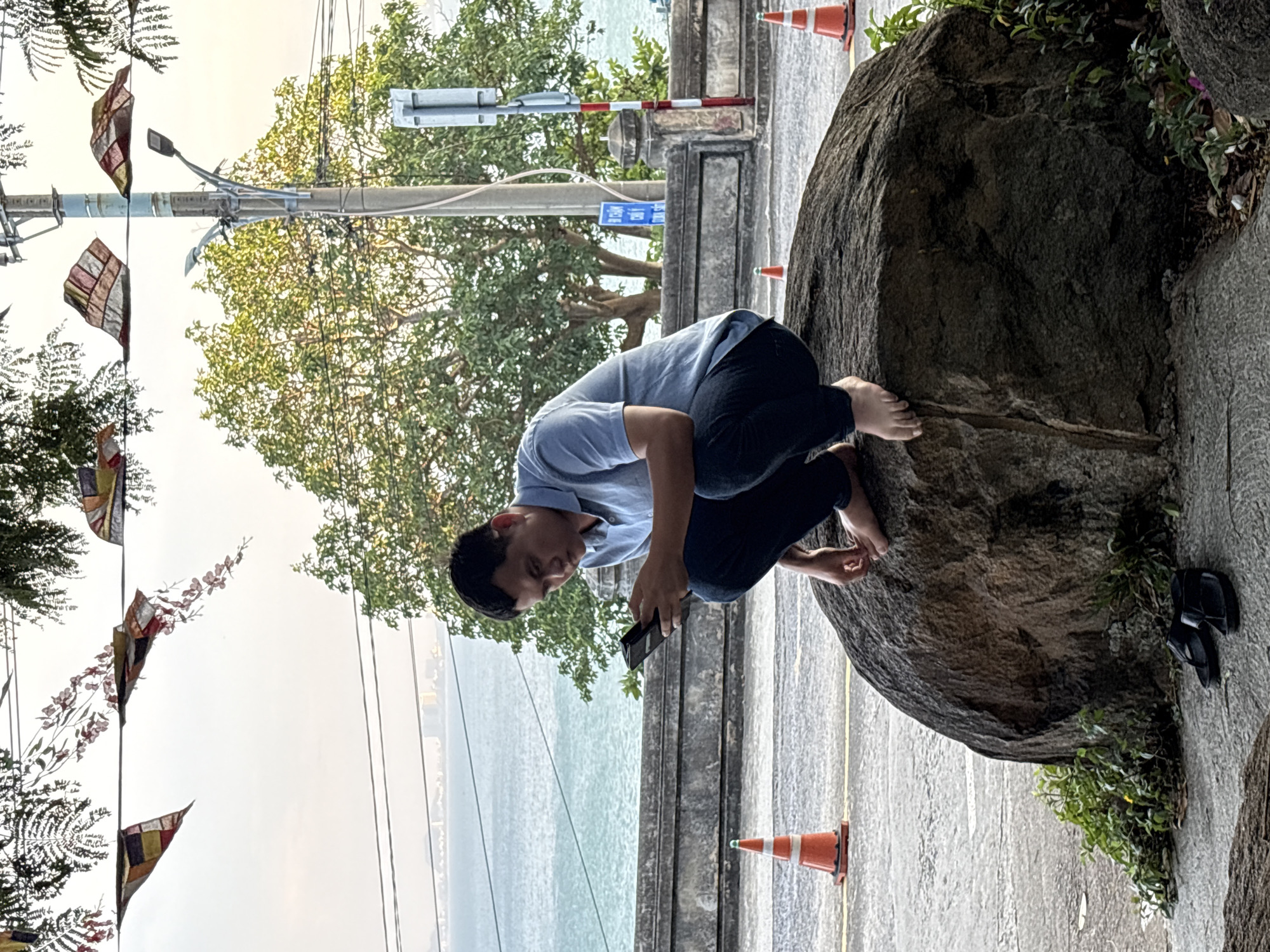 A man perches barefoot on a large rock next to a seaside promenade in Đà Nẵng, Vietnam. He is crouched, looking at a smartphone in his hands. He is wearing a light blue shirt and dark pants. Orange traffic cones are placed on the promenade. In the background is the ocean, and overhead are strung flags. A pair of sandals rests on the walkway beside the rock.