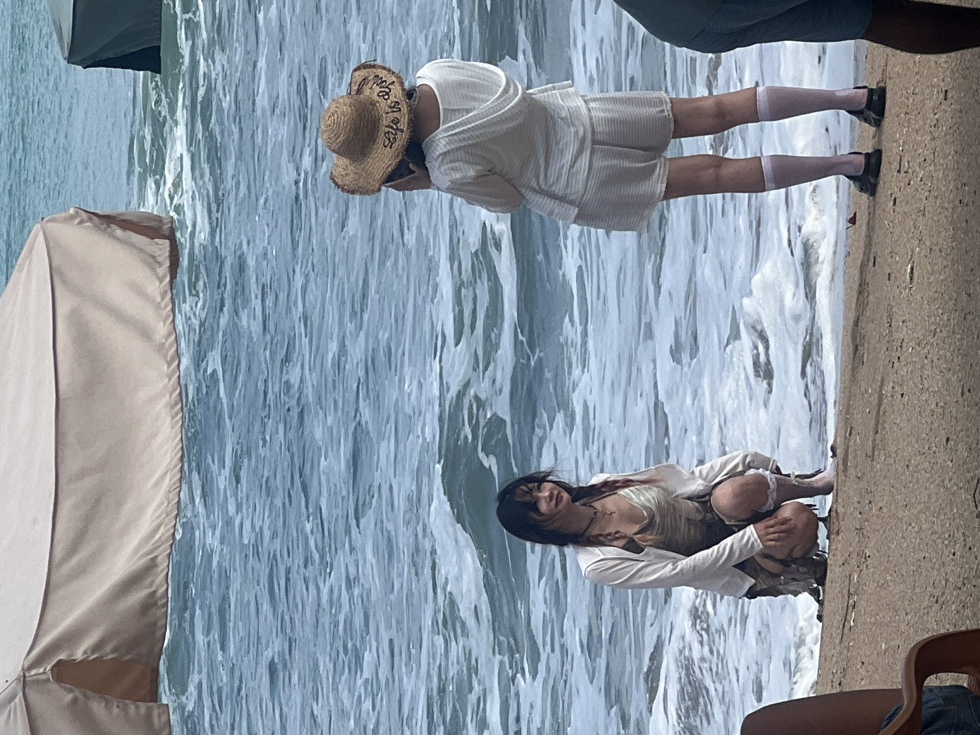 At the edge of a beach in Kuta, Indonesia, two people are present. One, a woman with dark hair in braids, crouches on the sand near the water. She wears a light-colored jacket over a patterned dress and white leg warmers. The second person, facing away from the camera, stands on the beach. They wear a straw hat with the text "Life is good" visible, a hooded top, and shorts, along with white socks and dark shoes. The ocean is visible behind them with waves breaking on the shore. A large, light-colored canopy is also partially visible in the background.