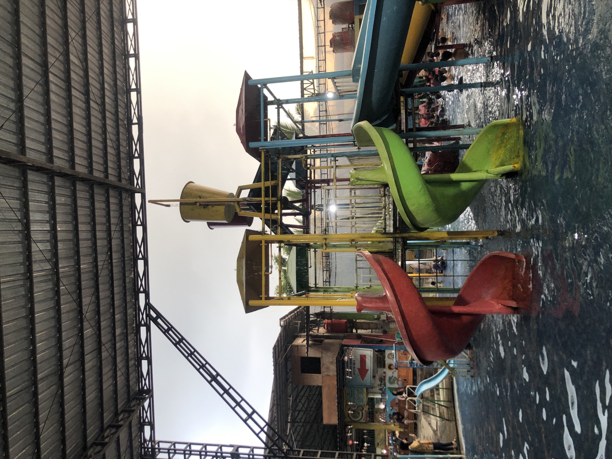 A wide-angle, eye-level shot captures a water park scene in Borobudur, Indonesia, under a large, corrugated metal roof supported by dark metal beams. The foreground is dominated by a shallow pool filled with dark, rippling water. Two prominent spiral water slides emerge from the pool: a vibrant red one on the left and a bright green one on the right.
Behind the slides, a multi-level water play structure is visible. It features yellow and blue support beams, yellow platforms, and a yellow bucket at the top, from which water appears to be falling. A green-roofed structure is also integrated into this play area. In the background, a two-story building with a brownish facade and a dark-tiled roof is situated. Signs, one with a red arrow pointing left, are affixed to the building.
Several people are present in the scene. In the pool, individuals are seen swimming or playing. On the left, near the building, a woman in a yellow top is taking a photograph with her phone, standing on the edge of the pool. Other people are seated on benches or standing near the building. The lighting suggests it is late afternoon, with sunlight filtering through the overcast sky and reflecting off the water and the metal roof. The overall atmosphere is one of a busy, recreational water park.