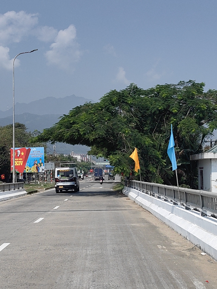 The image displays a daytime view of a multi-lane asphalt road, likely a bridge or overpass, in Hải Vân Ward, Vietnam. White solid and dashed lane markings are visible on the road surface. On the left, a tall grey street light pole stands adjacent to a large blue billboard featuring Vietnamese text, prominently "ĐẠI HỘI ĐẢNG BỘ ĐÀ NẴNG LẦN THỨ XIV," and illustrative graphics. A white van, bearing the word "KHANG" and a phone number on its rear, proceeds away from the viewer in the left lane. Further down the road, additional vehicles, including multiple trucks and motorcycles, are observed moving in the same direction. The right side of the road is bordered by a white concrete barrier topped with a grey metal railing. An orange flag and a light blue flag are mounted on poles affixed to this railing. Behind the flags and railing, a large tree with dense green foliage partially obscures a light-colored building with a green-tiled roof. In the background, undulating mountains are visible beneath a blue sky with scattered white clouds. The scene indicates active vehicular traffic on an urban or peri-urban thoroughfare.