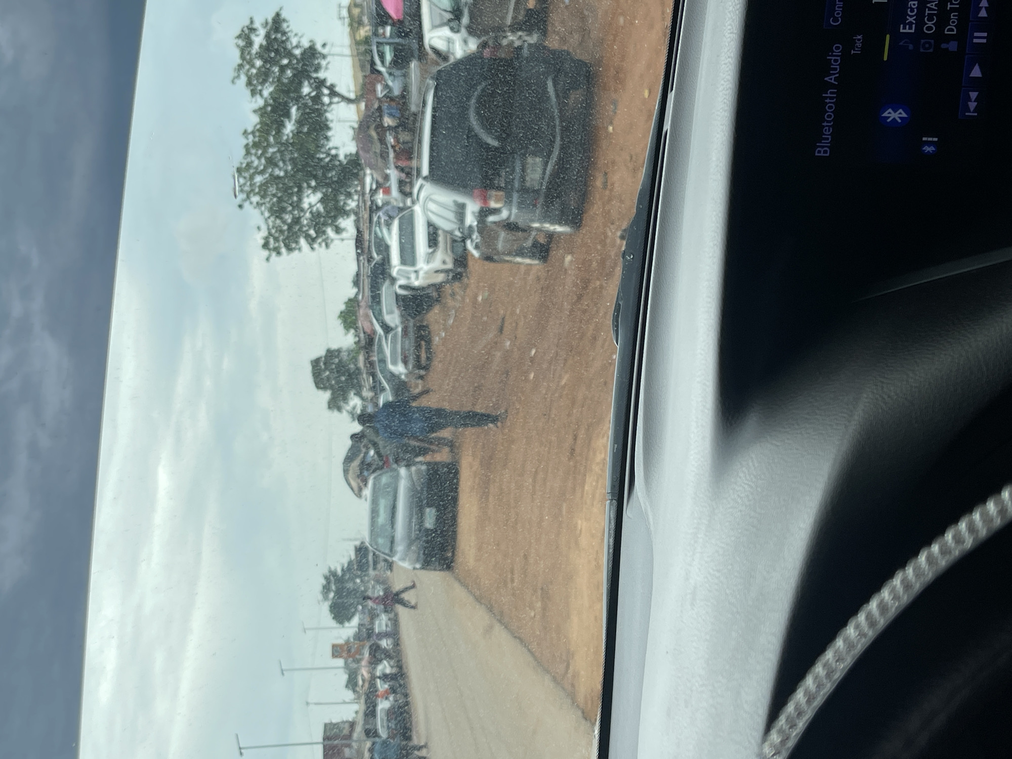 The image appears to have been taken from inside a vehicle in Karshi, Nigeria. It shows a dirt road crowded with parked cars and pedestrian traffic. A tree is visible in the background. A display screen of a car infotainment system is visible with “Bluetooth Audio” and audio track information.