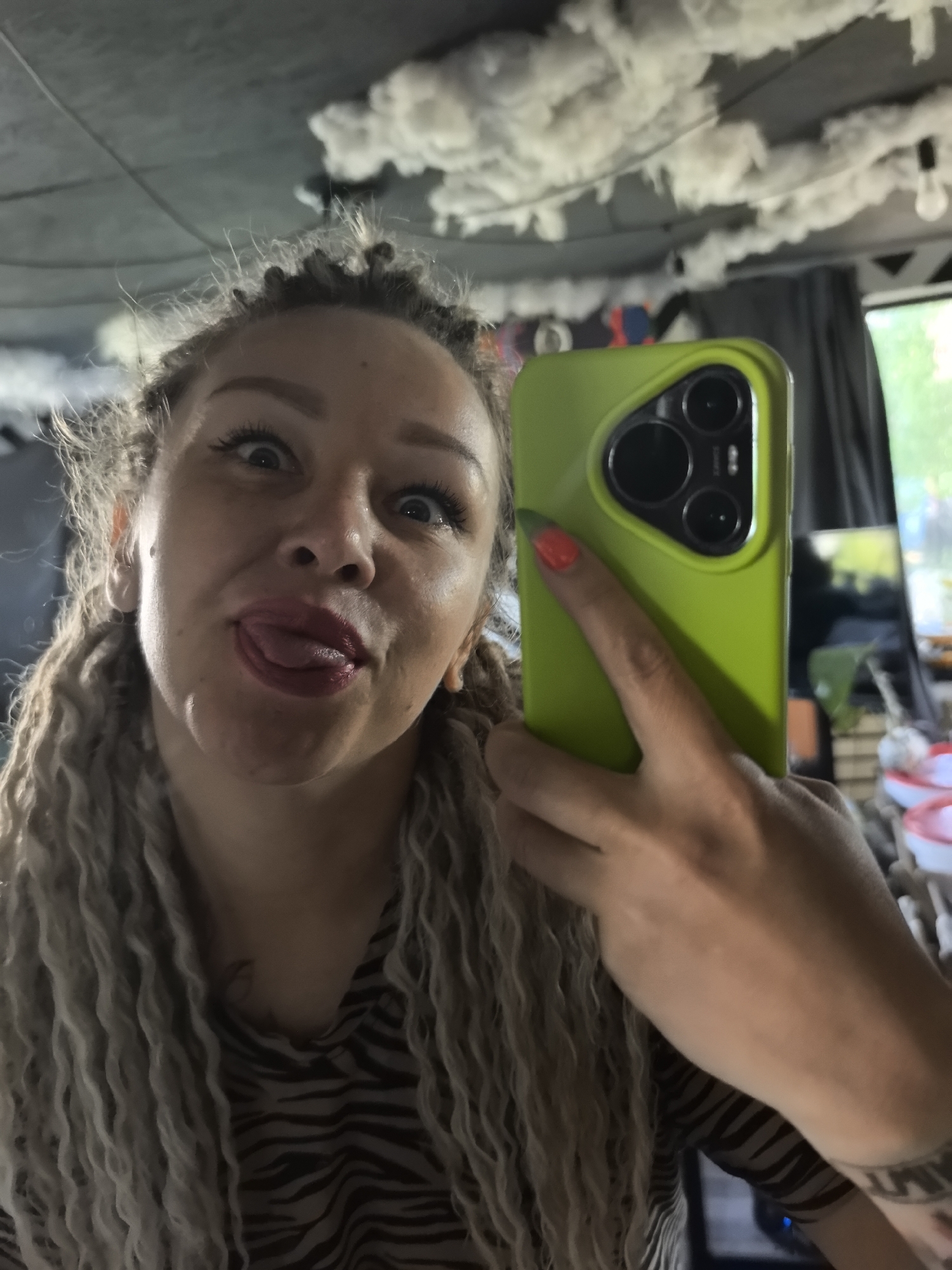 A woman with dreadlocks and a zebra-print shirt is taking a selfie with a green phone case. She is sticking her tongue out and making a silly face. The ceiling is decorated with what appears to be clouds made of cotton or a similar material. In the background, a window and some objects are visible, but details are unclear. The city of Noviy, Russia is not discernible from the image.
