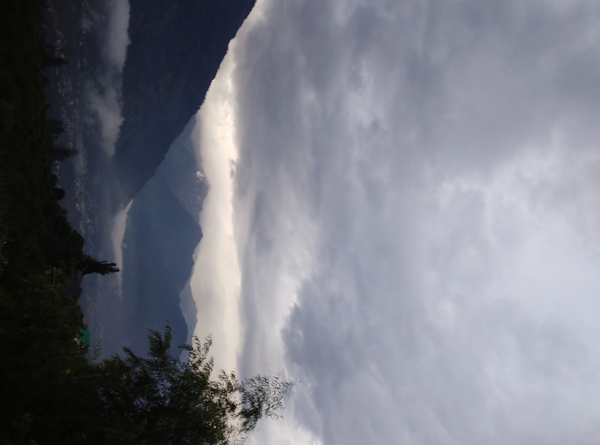 The image depicts a mountainous landscape under a cloudy sky. The foreground is dominated by dark foliage. In the mid-ground, a mountain slopes down towards a valley, with mist or low-lying clouds obscuring parts of a settlement below. Further in the background, several layers of mountains recede into the distance, with the peaks of some mountains visible above the clouds. The sky is filled with heavy, grey clouds, suggesting overcast weather or the approach of rain. A single tree with long, thin leaves is visible on the right side of the image.