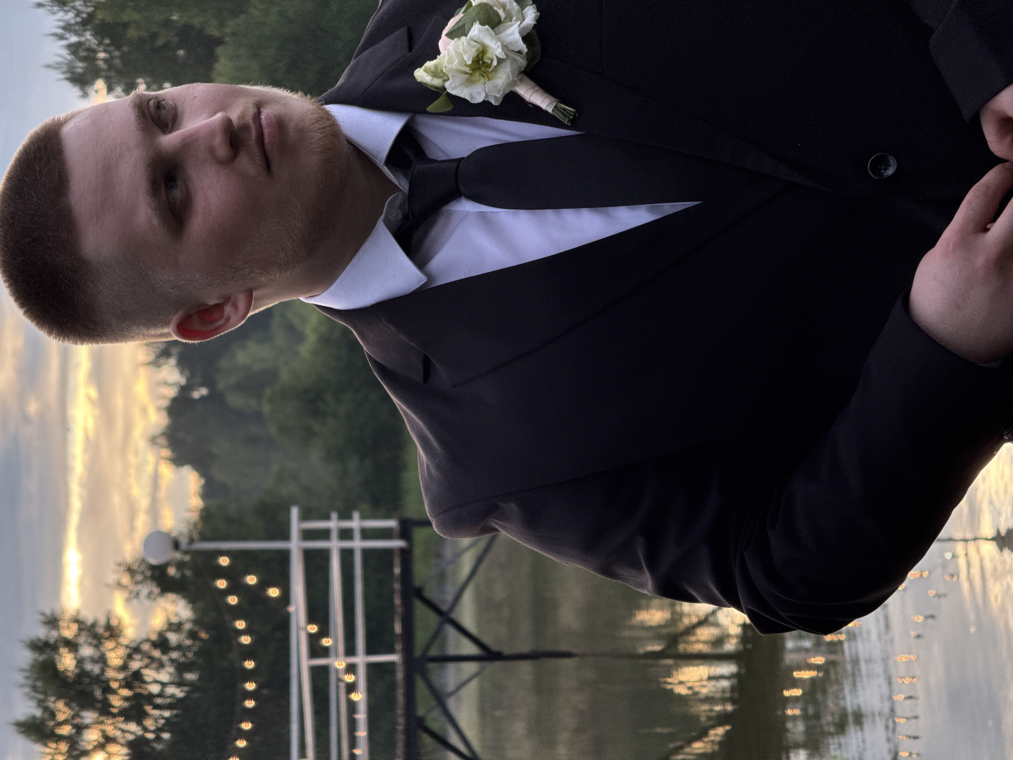 A medium shot shows a young man in a dark suit and tie, with a boutonniere on his left lapel. He has a buzz cut and light stubble. He is looking to his right, away from the camera. The background is blurred, but appears to be an outdoor setting with trees and a body of water. There are string lights visible in the distance. The lighting suggests it is dusk or early evening. The scene is likely Votkinsk, Russia.