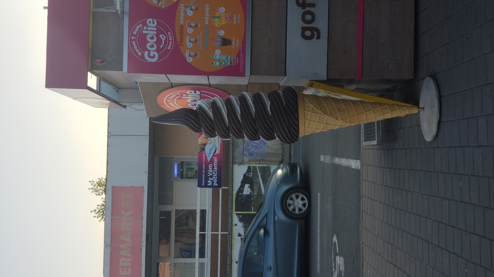This media file captures a street scene in Prešov, Slovakia, with a prominent, oversized sculpture of a chocolate and vanilla soft-serve ice cream cone as the central object. The sculpture is placed in front of a building housing a "Goolie" establishment, which appears to be a food or drink vendor, as indicated by the signage advertising "Horúce nápoje" (hot drinks) and "Studené nápoje" (cold drinks), along with images of coffee and bubble tea. Another sign visible on the building reads "SUPERMARKET."
A dark blue car is parked on the paved street, partially obscuring a sign that translates to "Didn't get a loan from the bank? We will lend you money!" This suggests the presence of a financial service or loan provider nearby. The overall setting appears to be an urban environment, possibly a commercial strip or parking lot, with a grey brick-patterned sidewalk and asphalt road.
The lighting suggests it is daytime, with a bright sky visible in the upper left portion of the image, indicating fair weather. The sculpture itself is a striking, playful element in the scene, drawing attention to the "Goolie" brand. The image has been rotated, so the signage and elements are presented vertically.