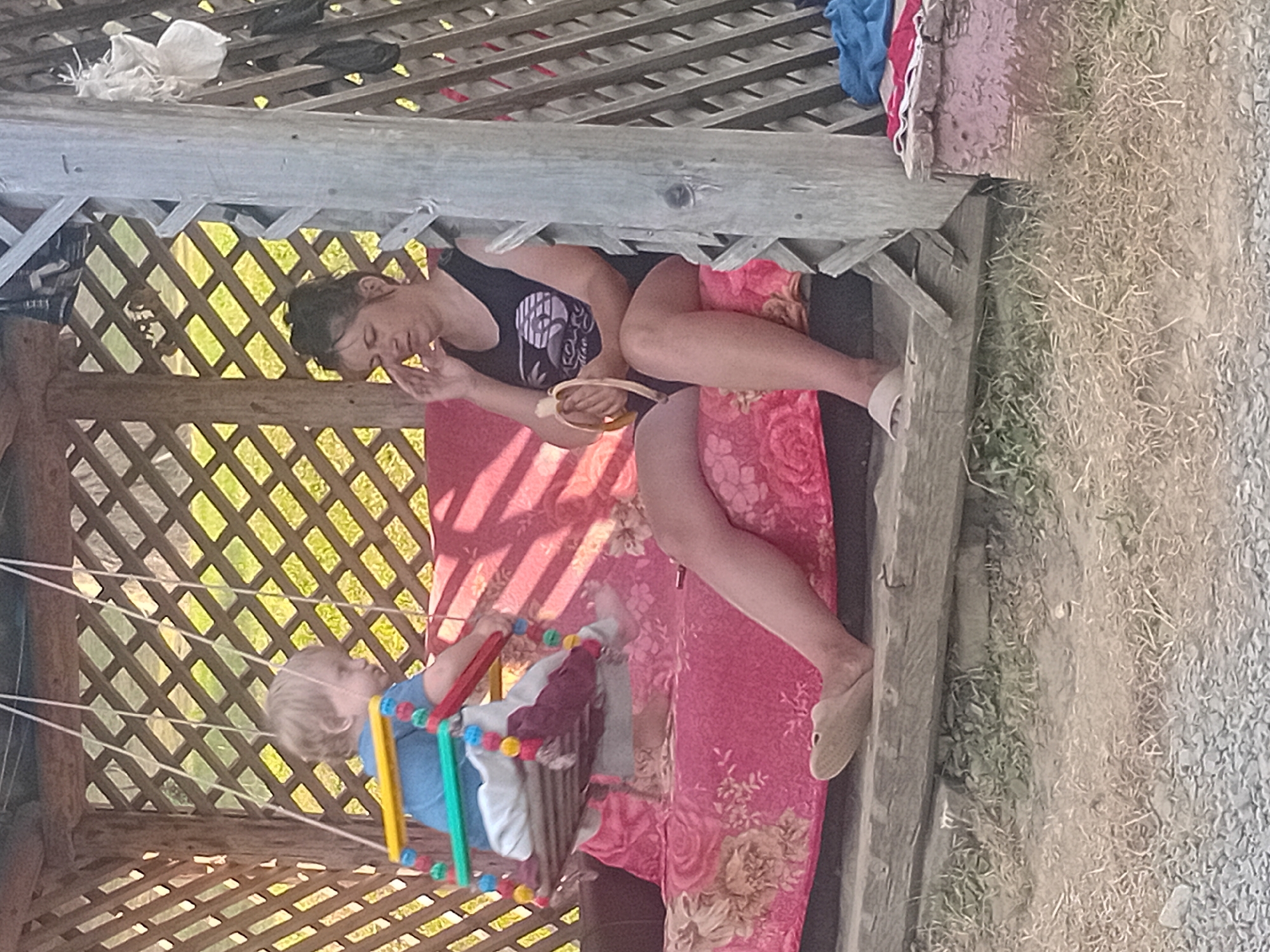 In this vertically oriented image, a young child is suspended in a colorful, homemade swing made of wooden rods and plastic beads, hanging from white ropes within a rustic wooden gazebo. The child, with blonde hair and wearing a blue shirt, appears to be a toddler and is looking to the side. To the right of the child, a woman reclines on a pink, floral-patterned cushion. She is wearing a black tank top and dark shorts, with her head tilted back and eyes closed, suggesting she is resting or napping. Her right hand is raised near her face, and she wears a light-colored shoe. The gazebo has a latticed wooden structure, allowing sunlight to filter through, casting striped shadows on the ground and the cushion. Behind the gazebo, a grassy area with some dry weeds is visible, and beyond that, gravel and more dry vegetation. The scene appears to be set outdoors during the daytime, likely in a rural or semi-rural location, given the natural surroundings and the simple construction of the gazebo. The overall mood is one of quiet relaxation and a peaceful moment outdoors. There is no visible text in the image.