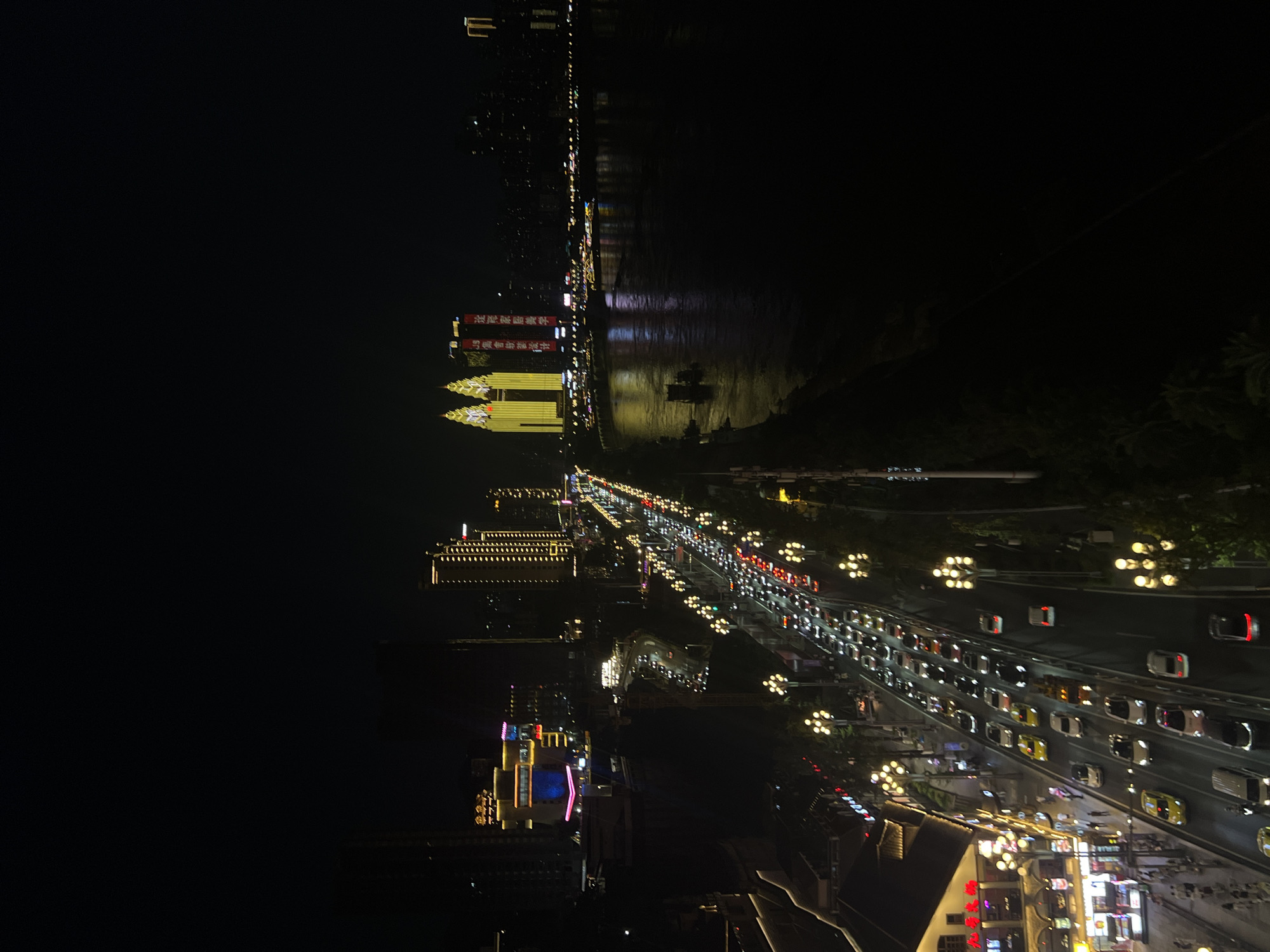 This is an aerial nighttime view of a busy waterfront street and river in Nan'an District, China. A multi-lane road is heavily trafficked with cars and trucks, their headlights and taillights creating streaks of light. On the left side of the road, there are buildings with illuminated signage and windows. Streetlights line the road, some with decorative lamp posts. Across the road, a wide river reflects the lights from the city. On the far side of the river, several prominent skyscrapers are illuminated, including two distinctive twin towers with bright yellow facades. Boats are visible on the river. The overall scene depicts a vibrant urban night environment.